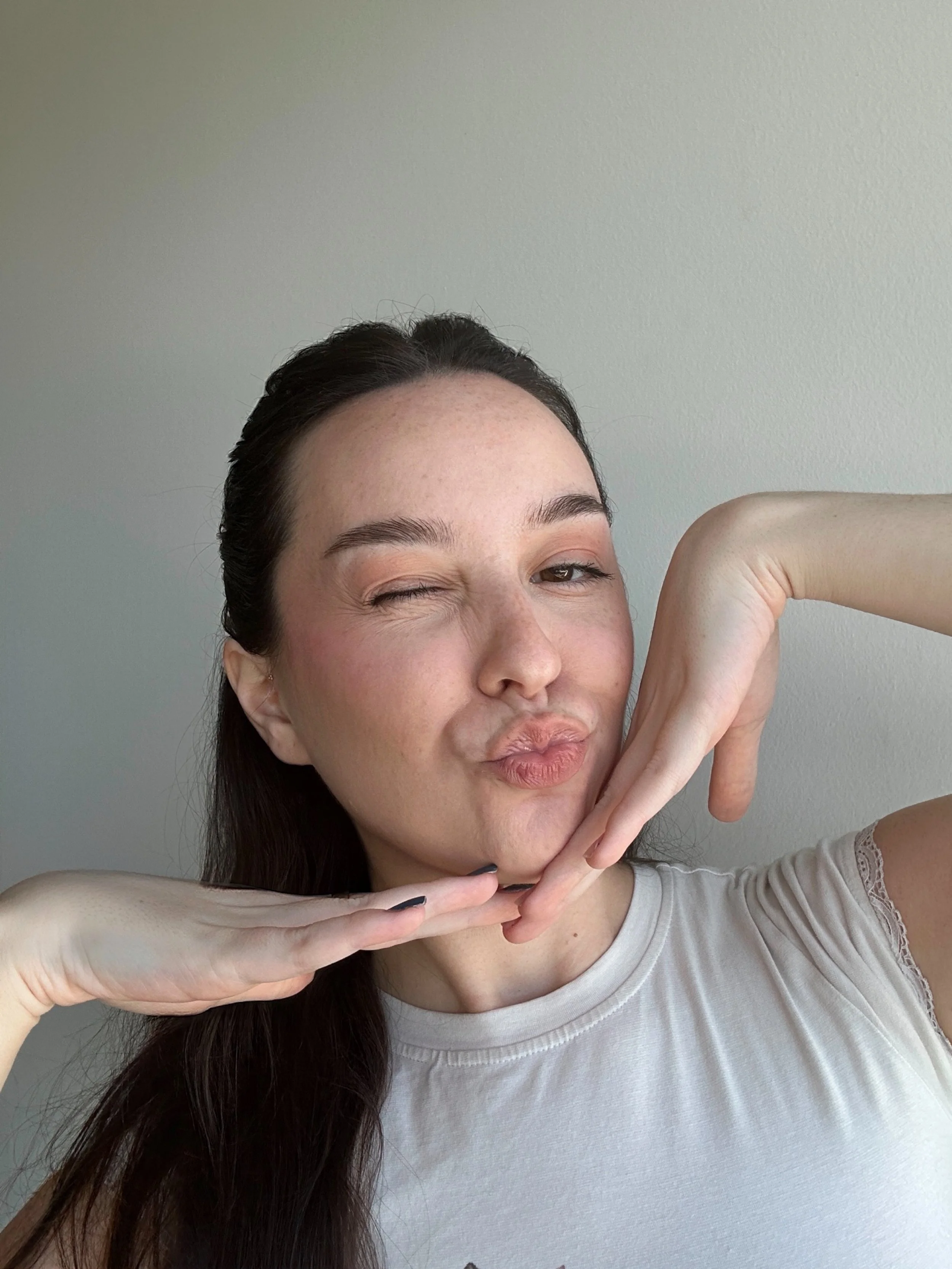 A woman with long dark hair making a playful pose with her left eye winking and her lips puckered, touching her chin with her left hand and her right hand near her face, against a plain light-colored wall.