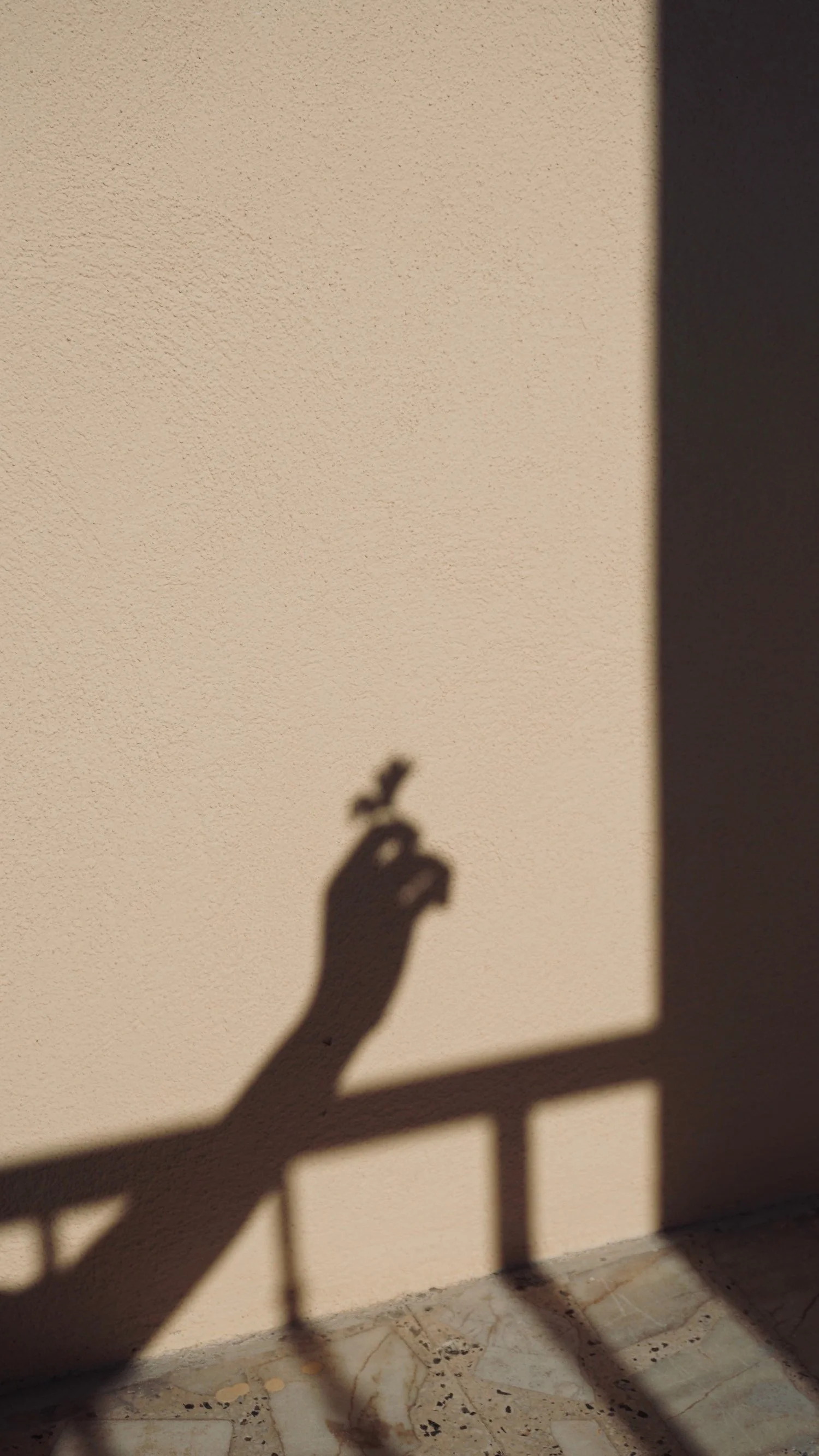 Shadow of a hand holding a cigarette cast on a beige wall with a railing and marble floor.