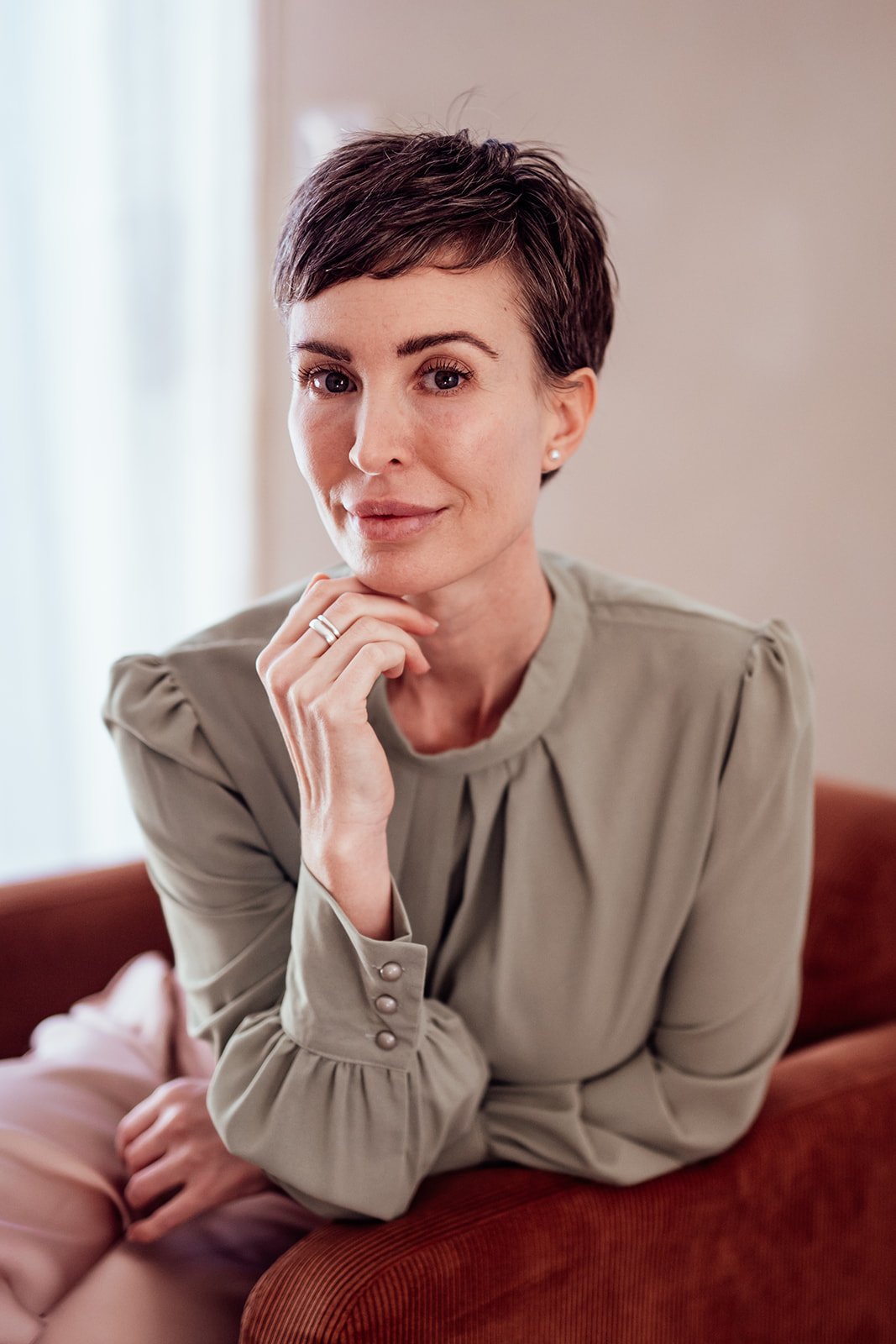 A woman with short dark hair and light skin, sitting on a red couch, resting her chin on her hand and looking at the camera.