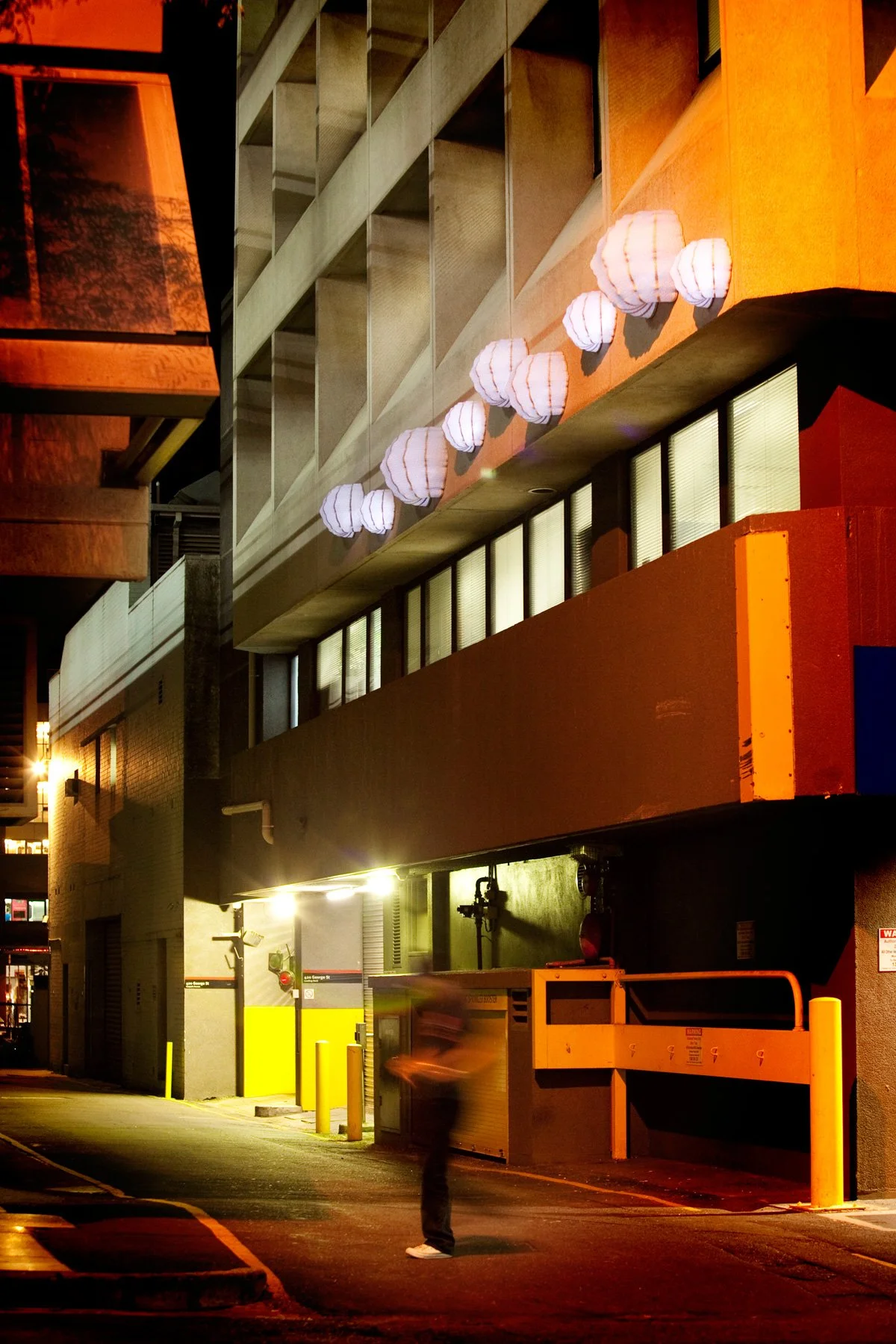 Lanterns by KT Doyle illuminated above Briggs Lane at night, with a pedestrian passing beneath the installation