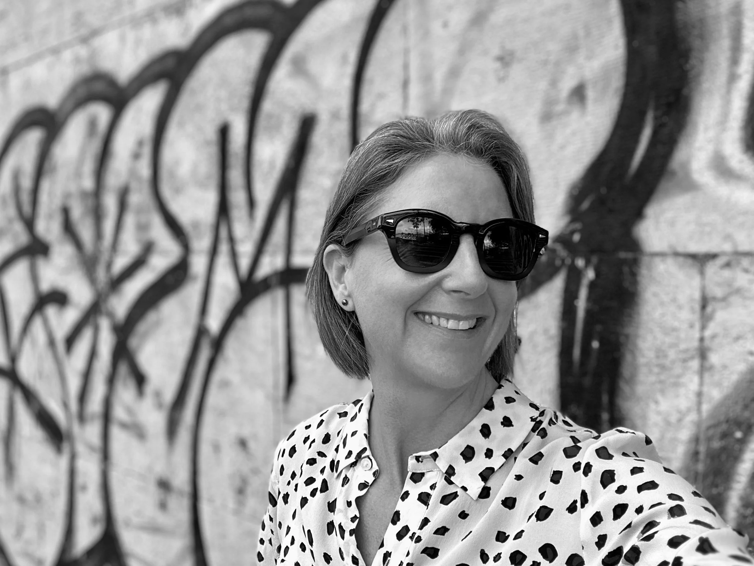 A woman smiling in front of a graffiti wall, wearing sunglasses and a polka dot shirt.