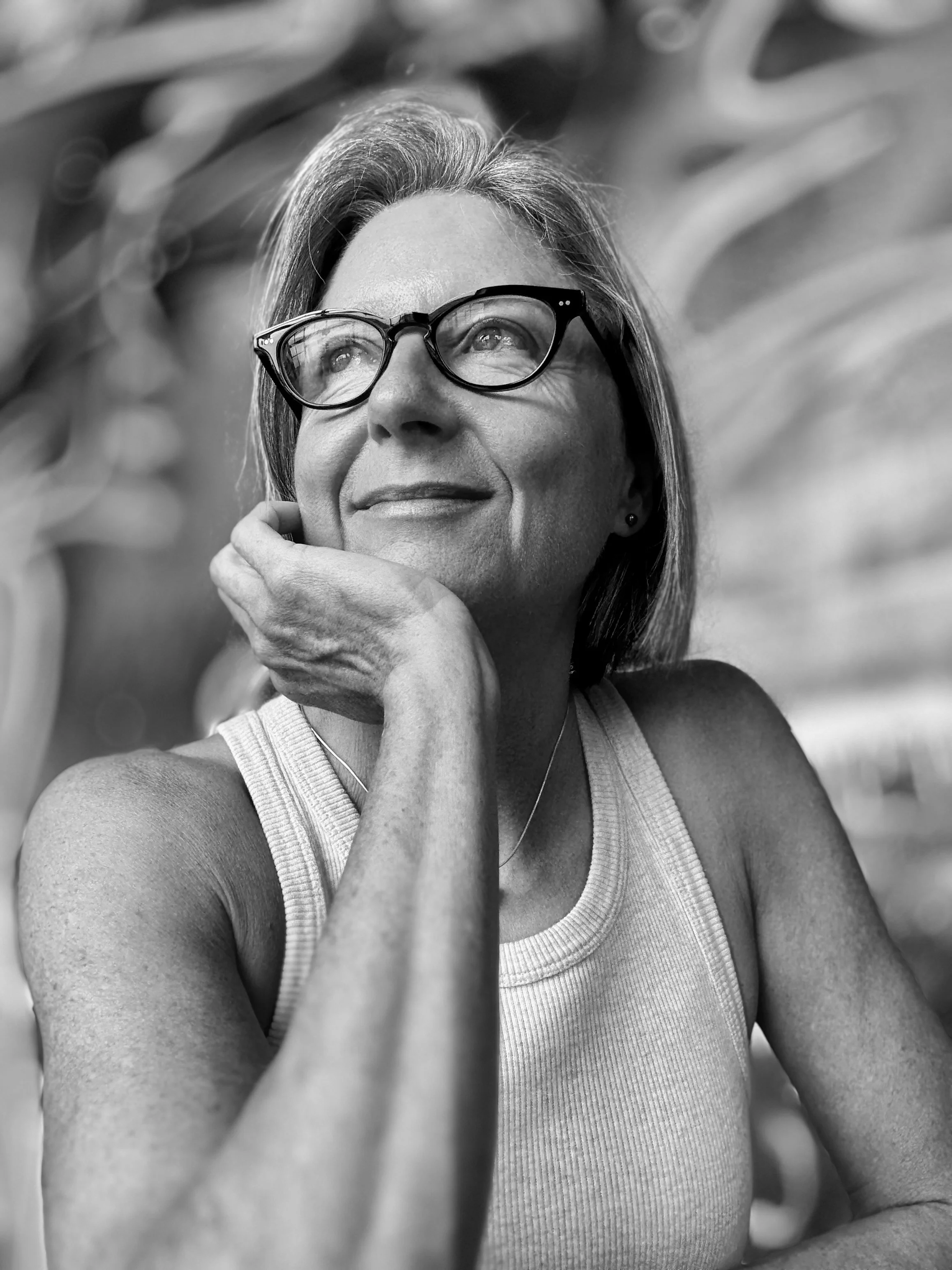 A woman wearing glasses and a sleeveless top, resting her chin on her hand, looking thoughtfully upward, with a blurred outdoor background.