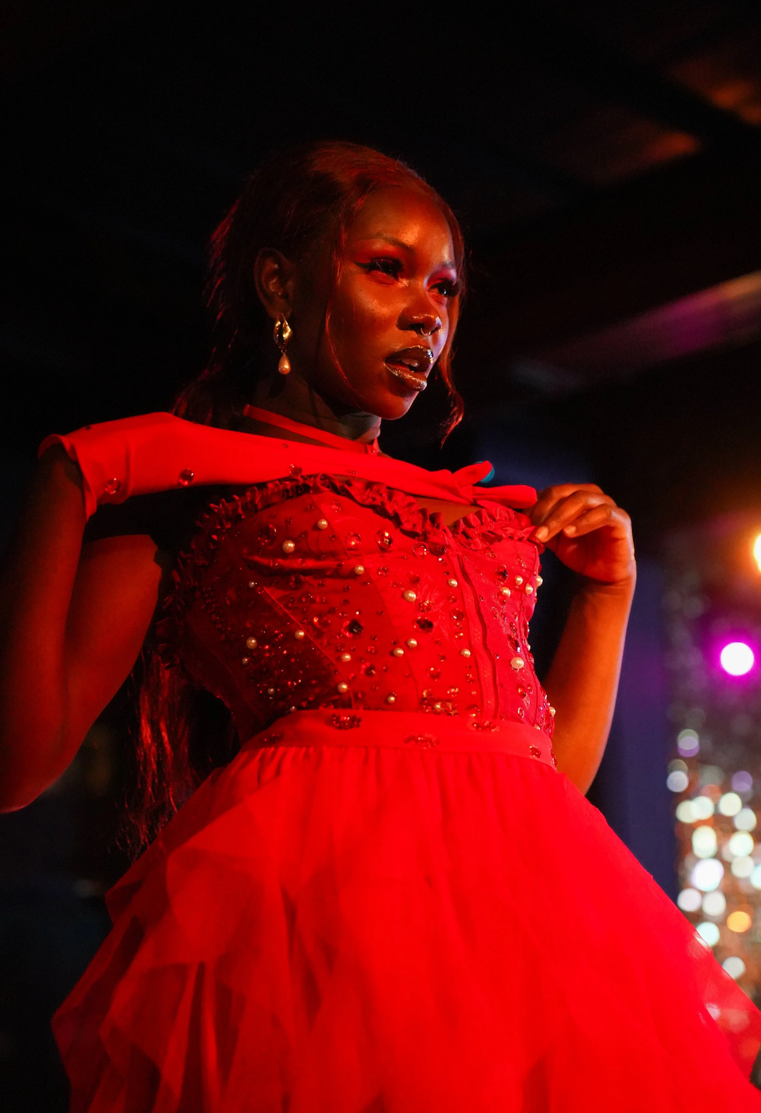 A woman with dark skin wearing a red embellished dress with ruffles, pearl details, and a bow, and pearl earrings, is presented against a dark background with blurred city lights.