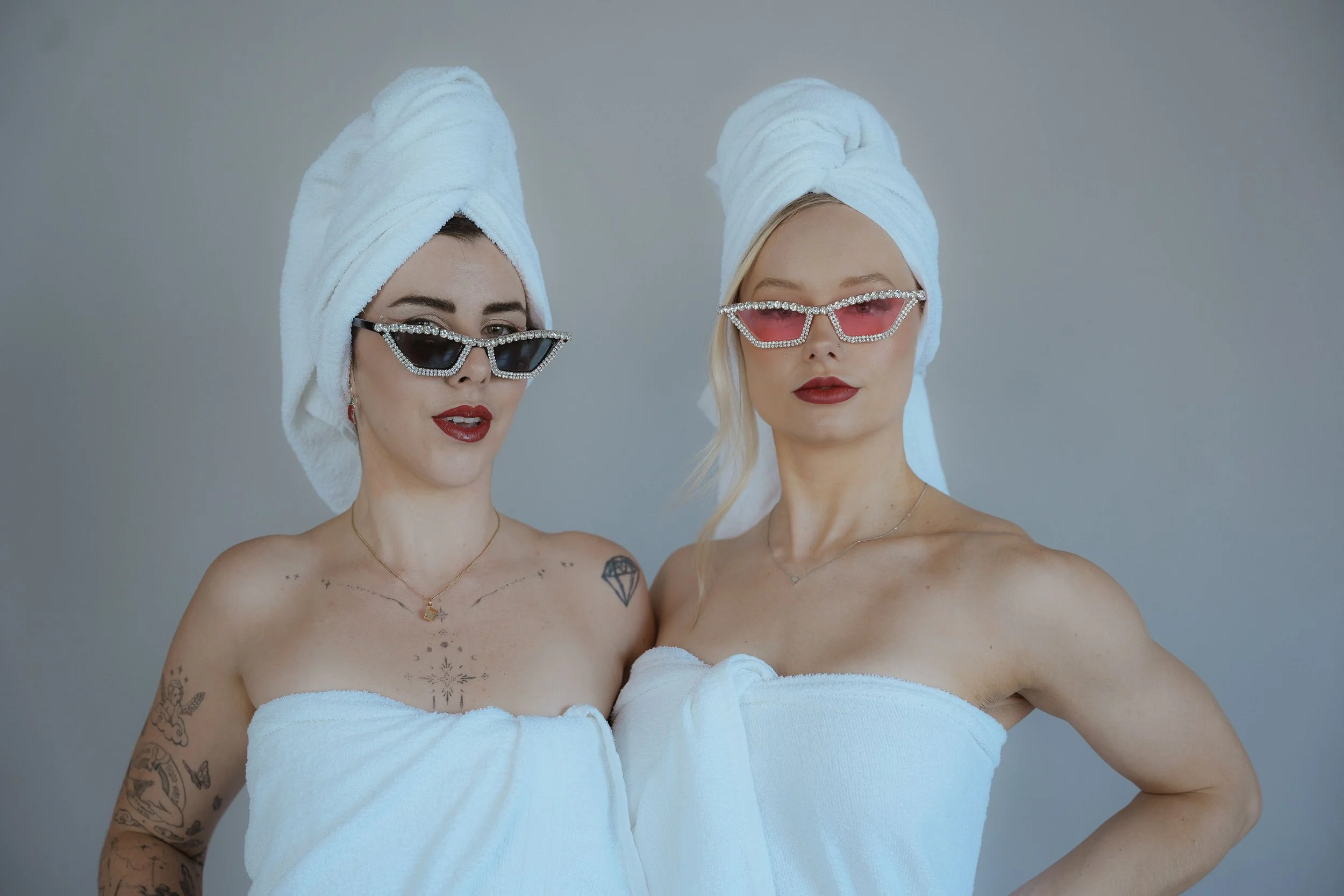 Two women wrapped in white towels with towels on their heads and wearing sunglasses, standing shirtless against a gray background.