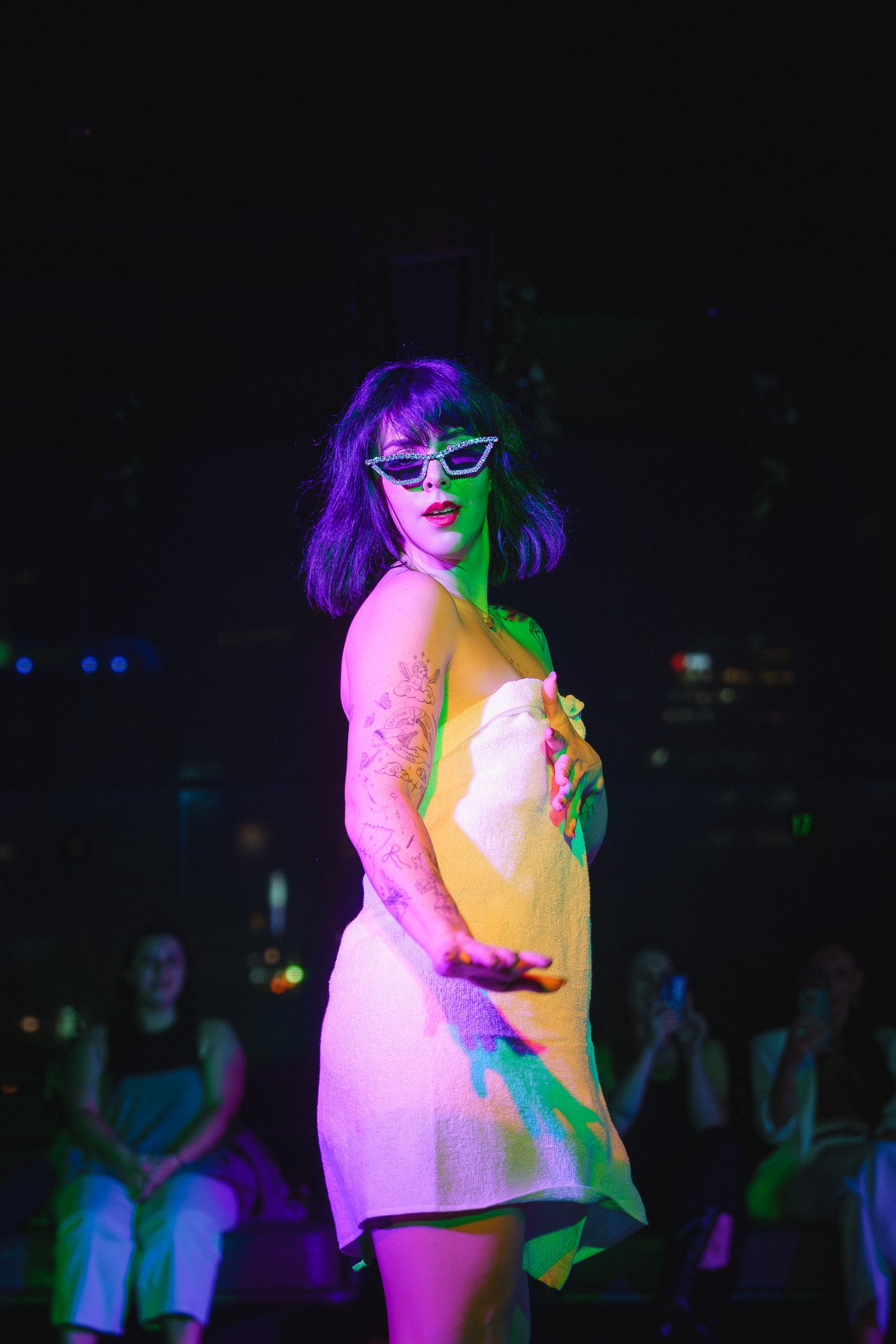 Gigi Cartier wearing sunglasses and a white towel is performing on stage at night, with several people sitting in the background.