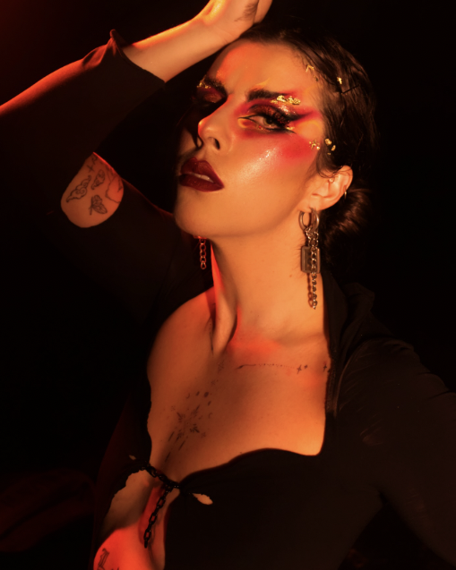 A woman with dramatic makeup, gold glitter, and styled dark hair, posing with one hand on her forehead against a dark background.