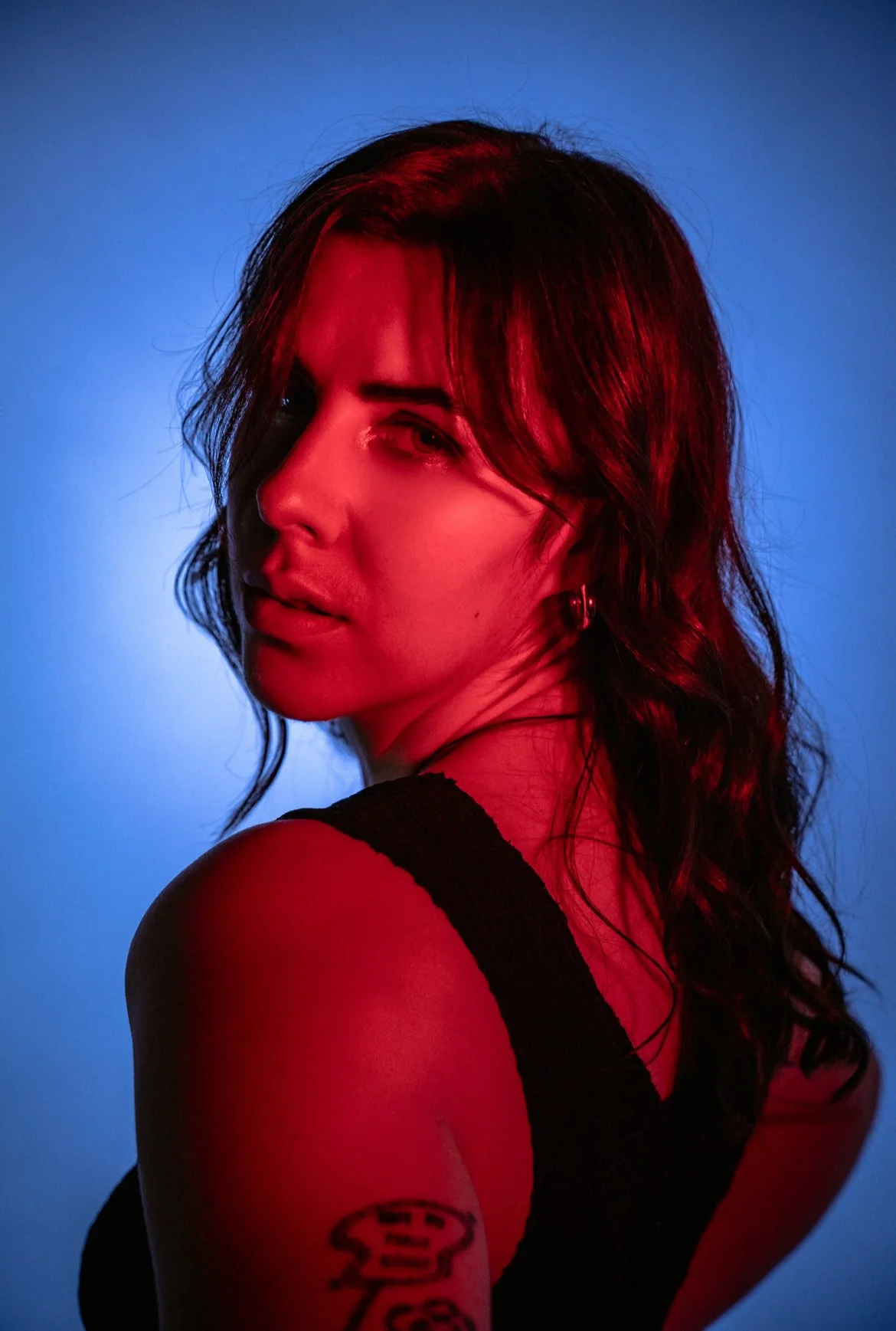 A woman with dark hair and earrings, looking over her shoulder, illuminated by red and blue lighting.