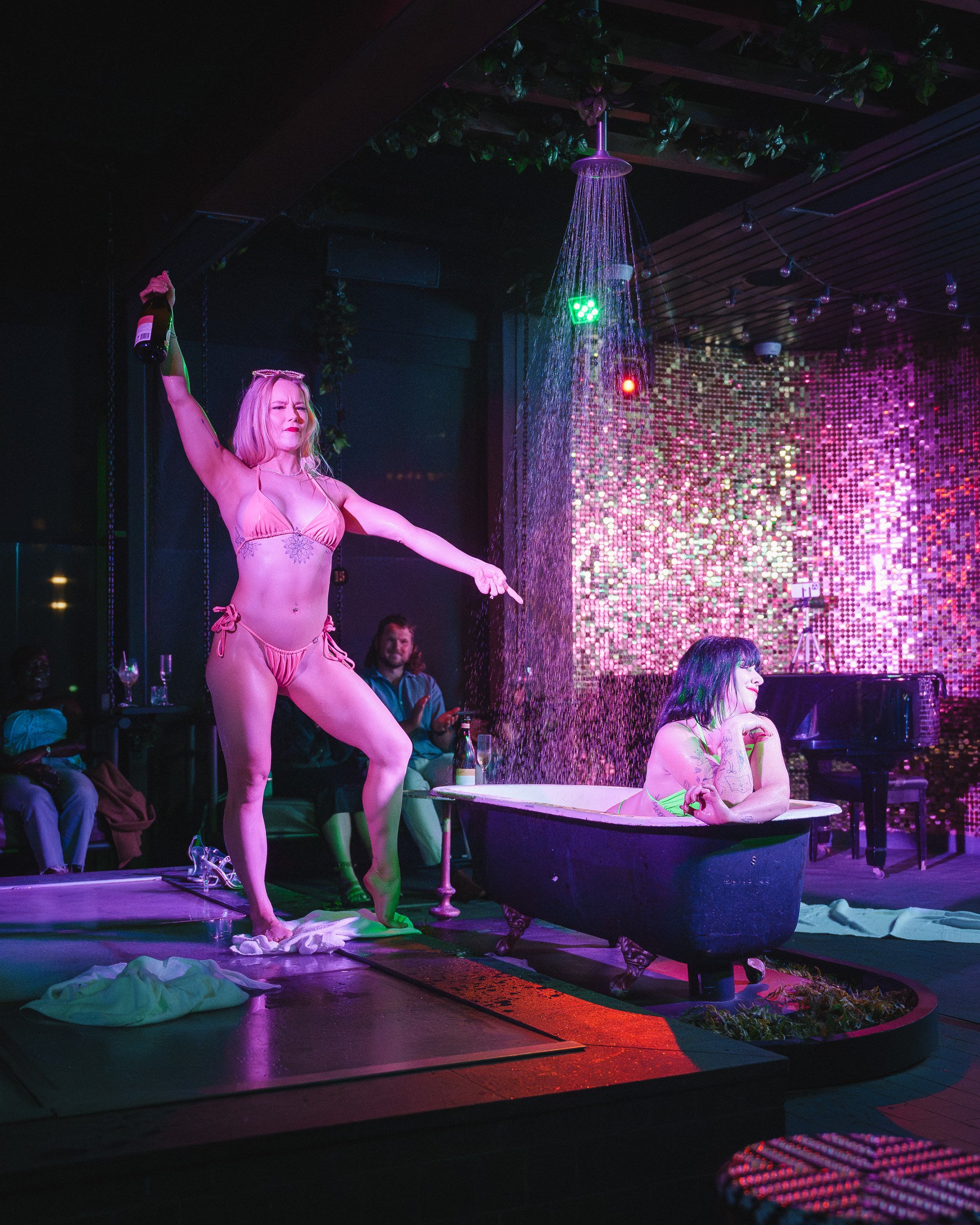 A woman sits in a bathtub on a stage with an embellished or reflective wall behind her. Another woman stands next to her, holding an object resembling a bottle or container, while wearing a pink bikini. The scene is illuminated with colorful stage lights, creating a vibrant and theatrical atmosphere.