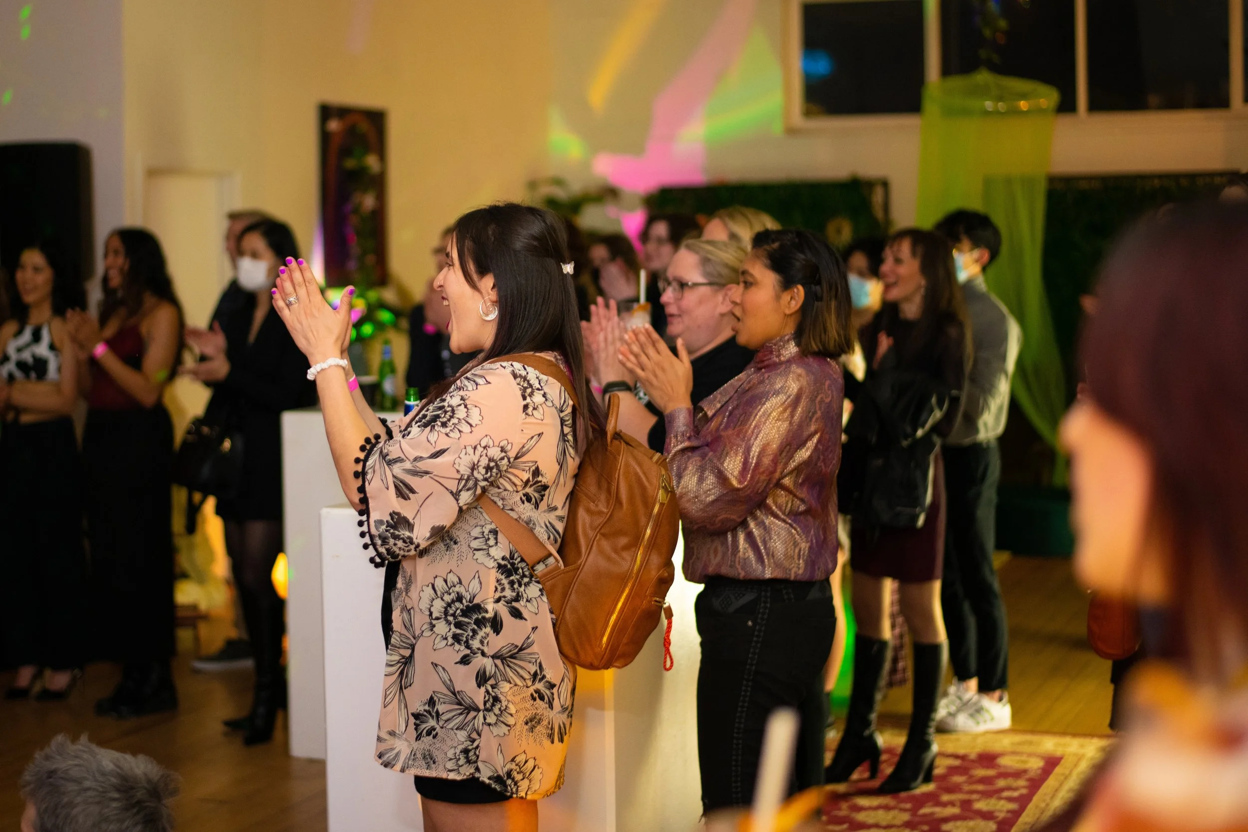 People gathered at an indoor event, some with hands raised in applause or prayer. The focus is on a woman in the foreground wearing a floral dress and jewelry, with others in the background. The setting includes colorful lighting and decorations.
