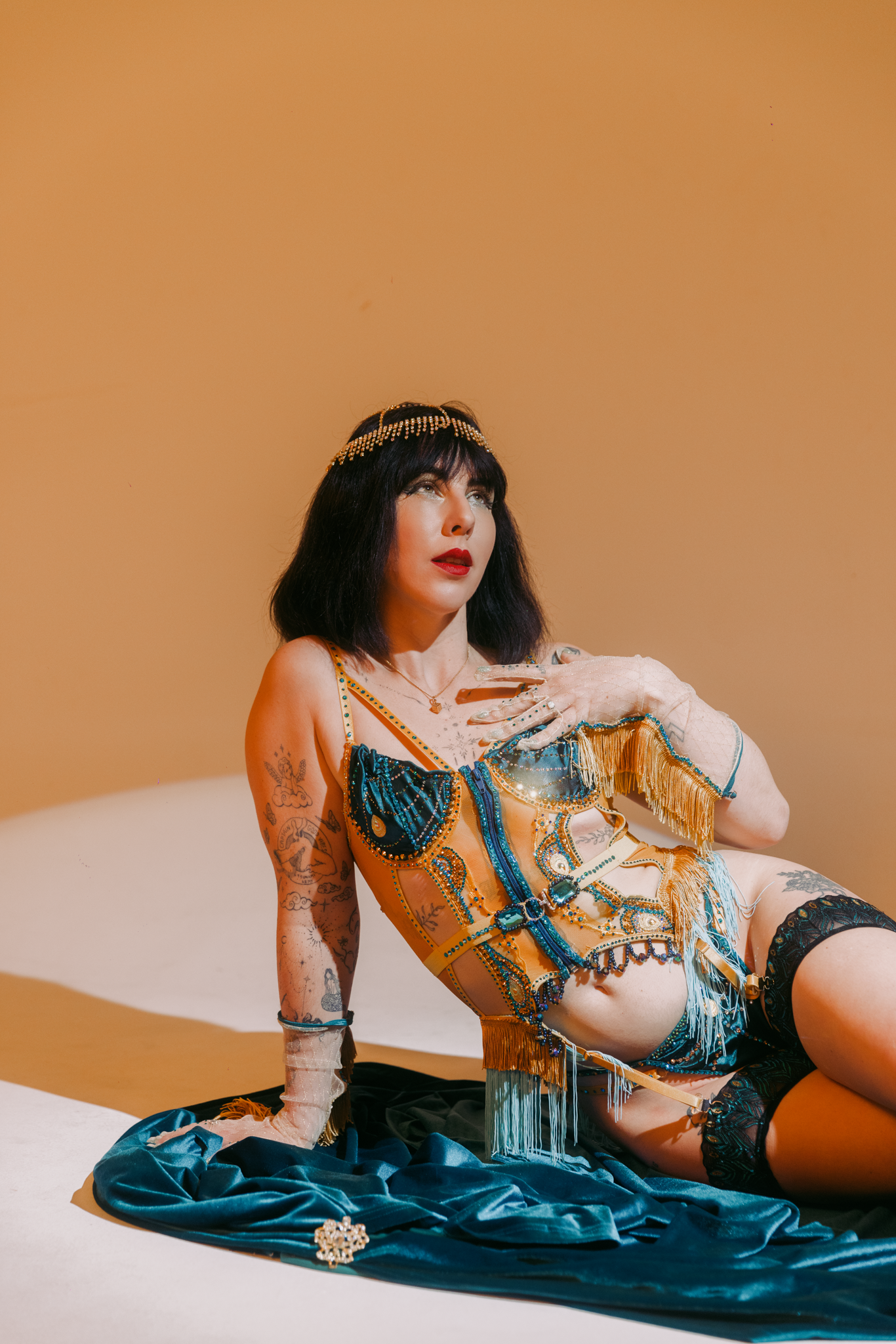 A woman with black hair and tattoos on her arm, dressed in a colorful, beaded, and fringed outfit, is sitting on the floor with a blue velvet material underneath her, against a plain, peach-colored background.