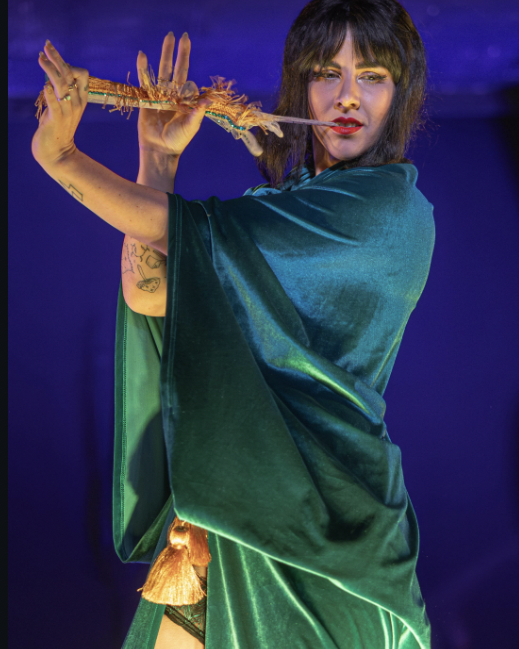 A woman with dark hair and tattoos on her arm holding her glove between her teeth and fingers as if she is playing a flute, wearing a velvet teal dress, against a dark blue background.