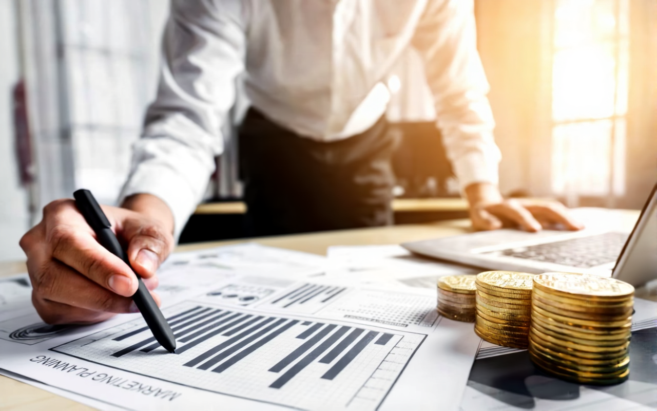 A person in business attire analyzing financial charts with stacks of gold coins on a desk, using a pen and a laptop.