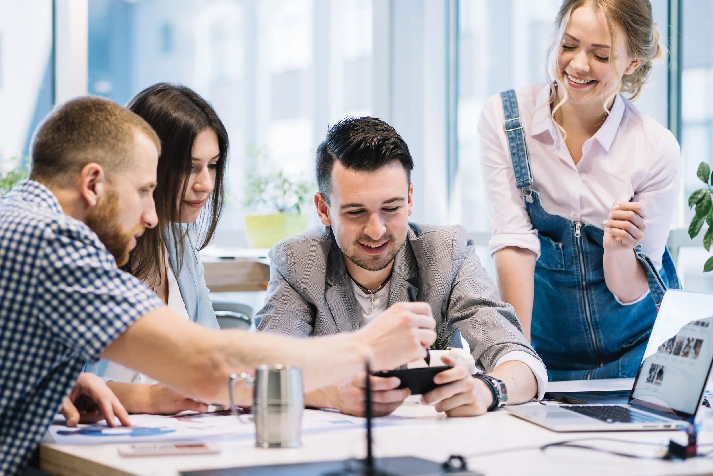Five young adults gathered around a table, looking at a smartphone and smiling, in a bright modern office.
