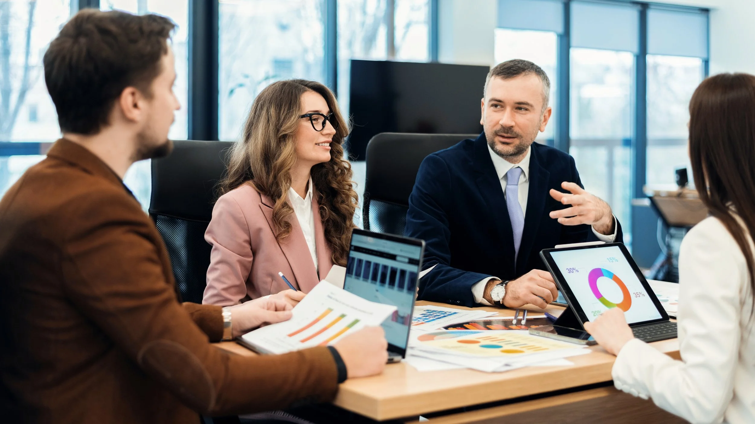 Business meeting with four people sitting around a table, discussing data on charts and tablets in a modern conference room.