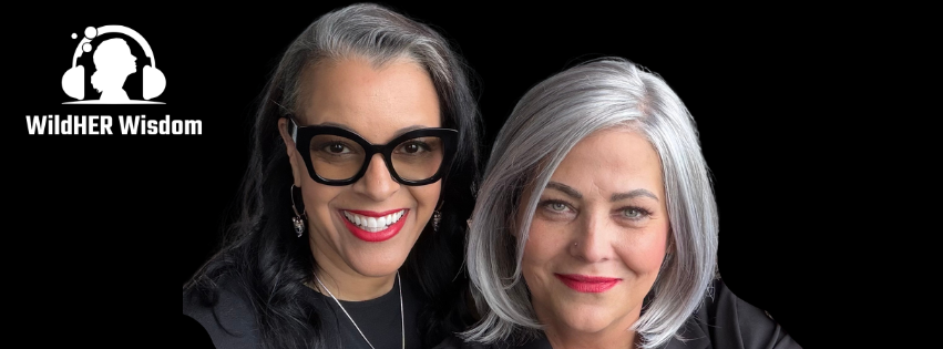 Two smiling women with gray hair, one with black glasses, dark clothing, and red lipstick, posing together against a black background. A logo with headphones and the text 'WildHER Wisdom' is in the top left corner.