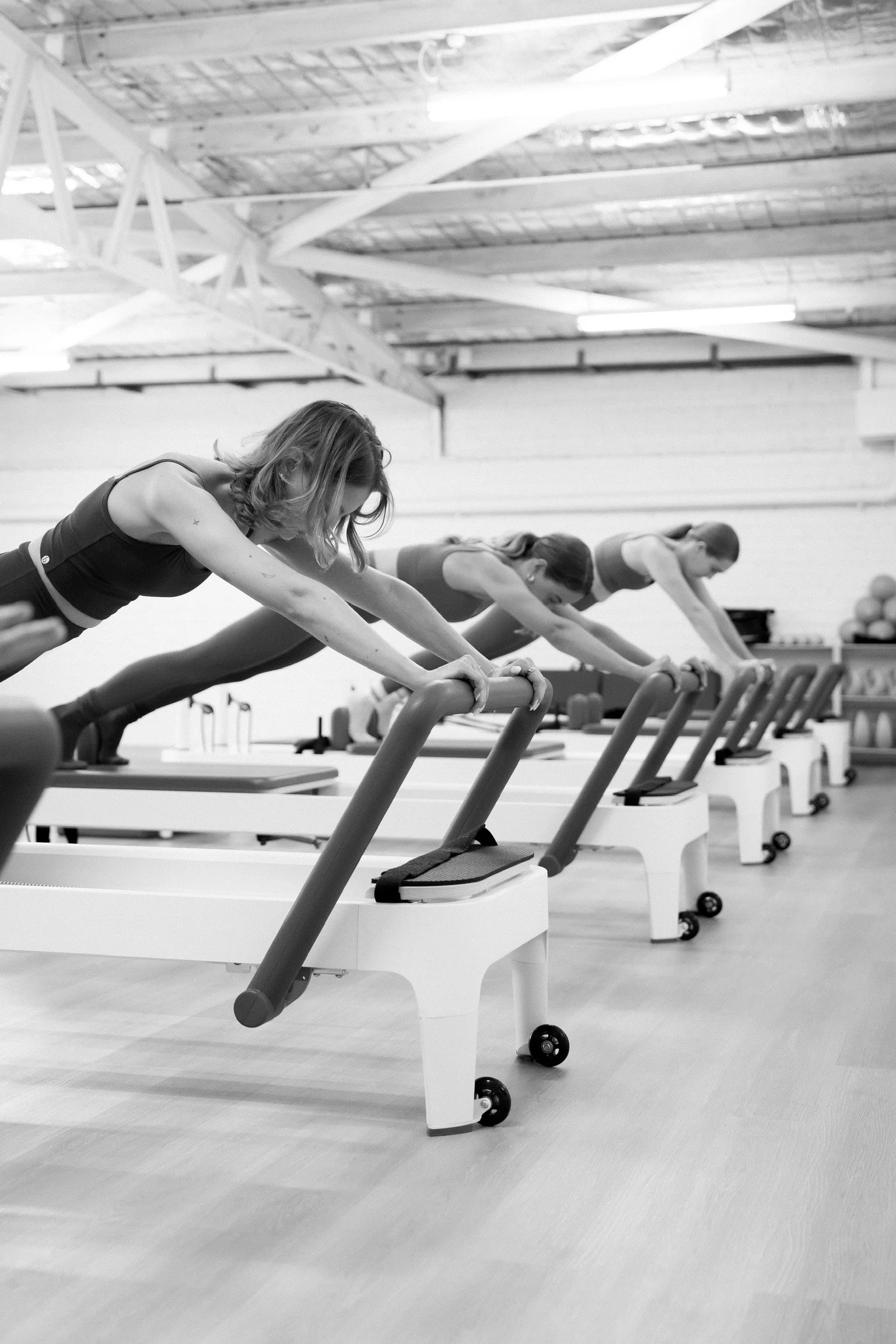Black and white photo of three women exercising on Pilates reformer machines in a fitness studio with a high ceiling and exposed beams.