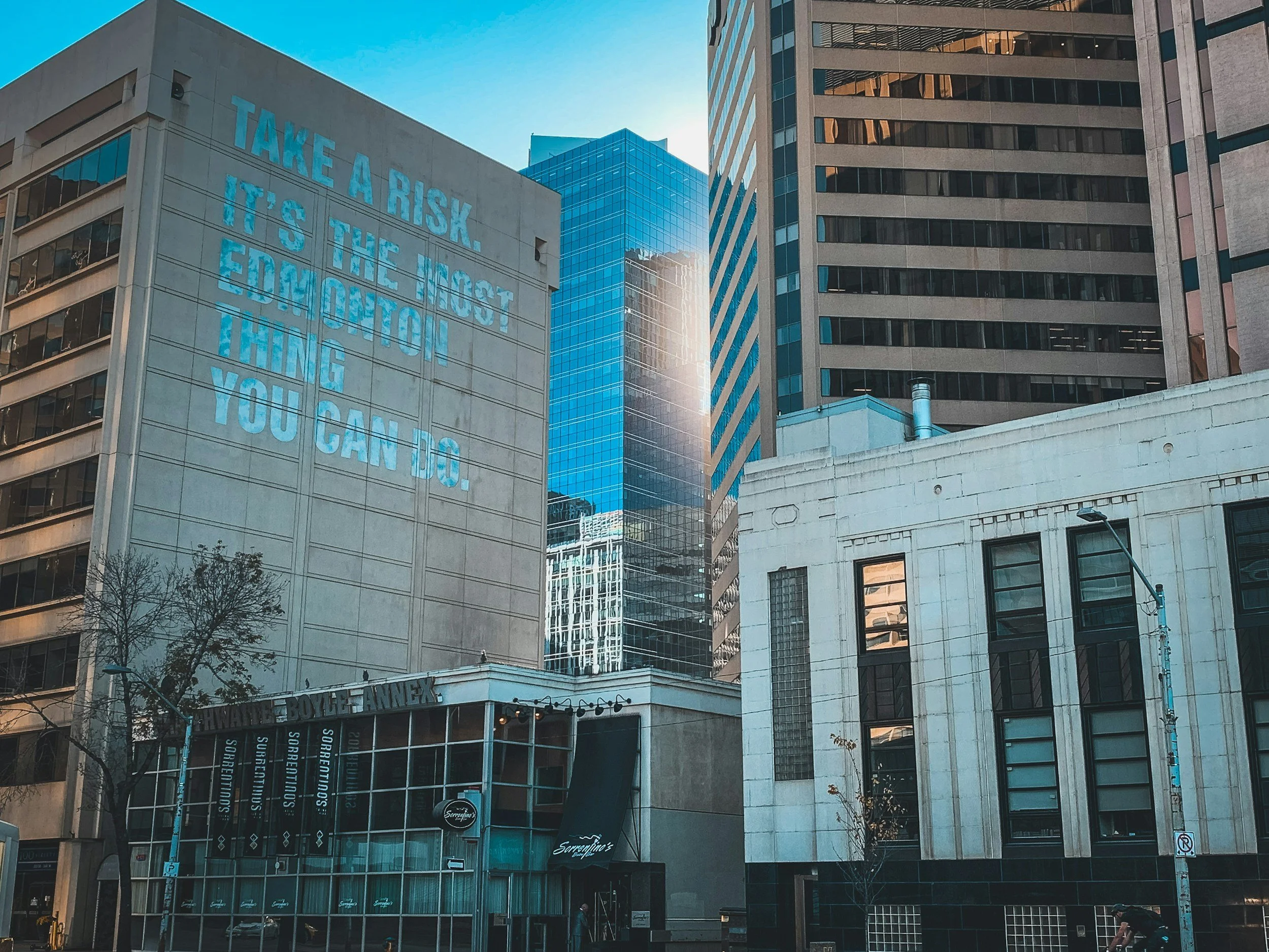 Cityscape with tall office buildings, one displaying a large blue neon sign with the message "Take a risk. It's the most education thing you can do."