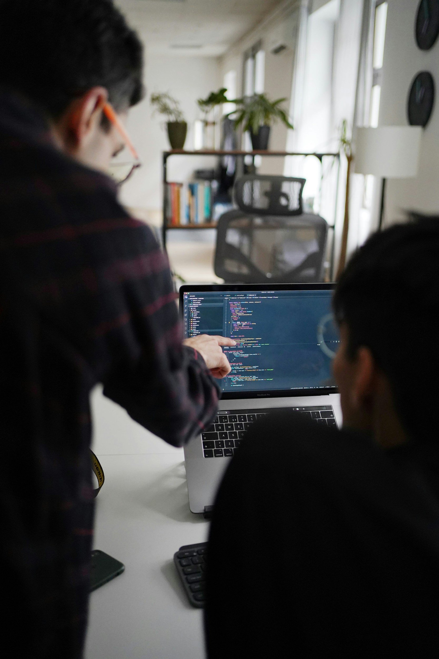Two people working on a computer with a screen displaying code in a bright room with shelves and plants in the background.