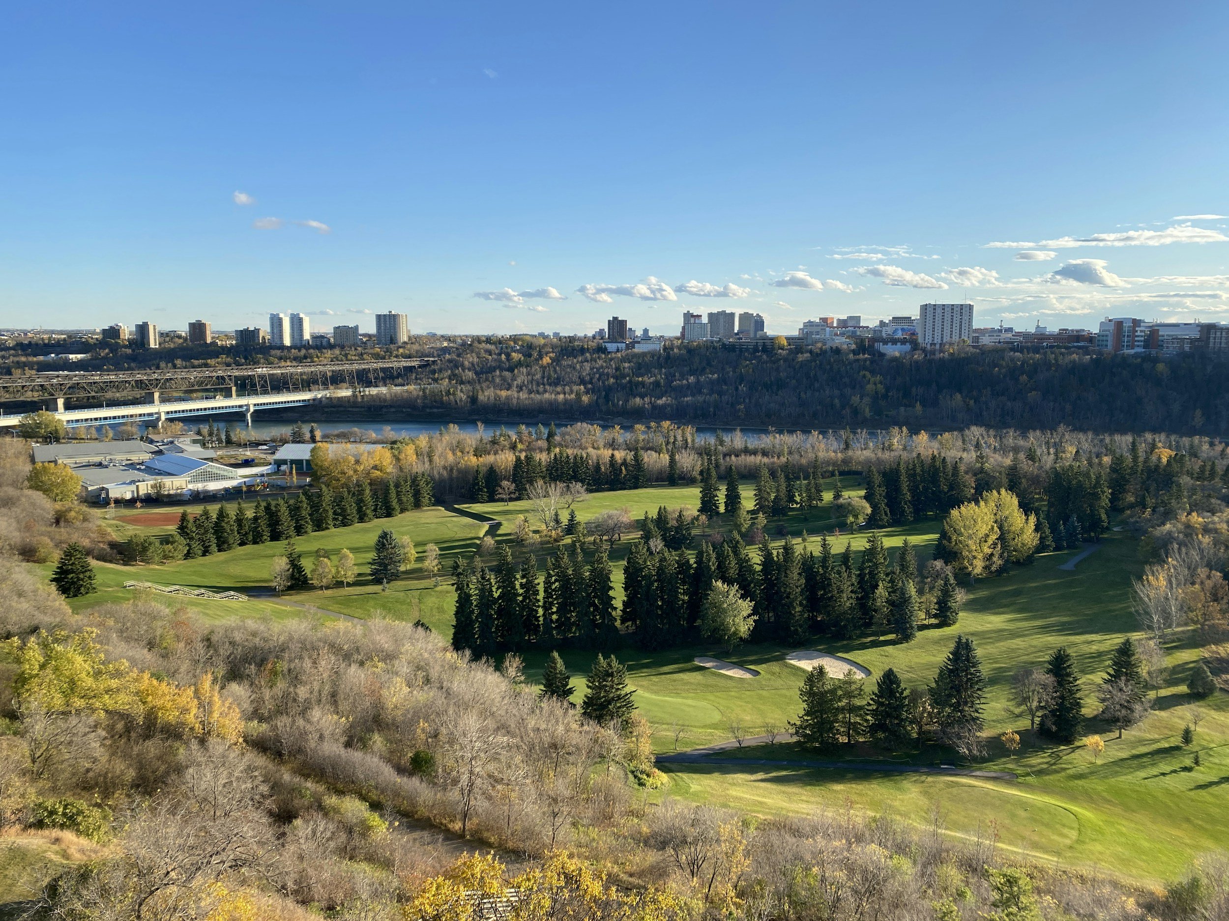 A scenic view of a golf course surrounded by trees with a cityscape and river in the background under a clear blue sky.