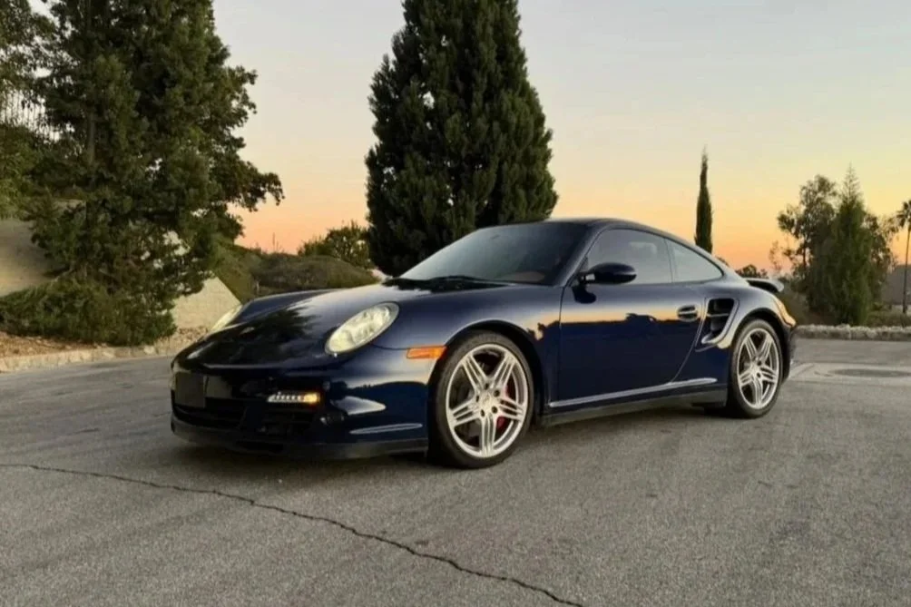 A dark blue Porsche sports car parked on an empty paved lot during sunset, with trees and a soft sky in the background.