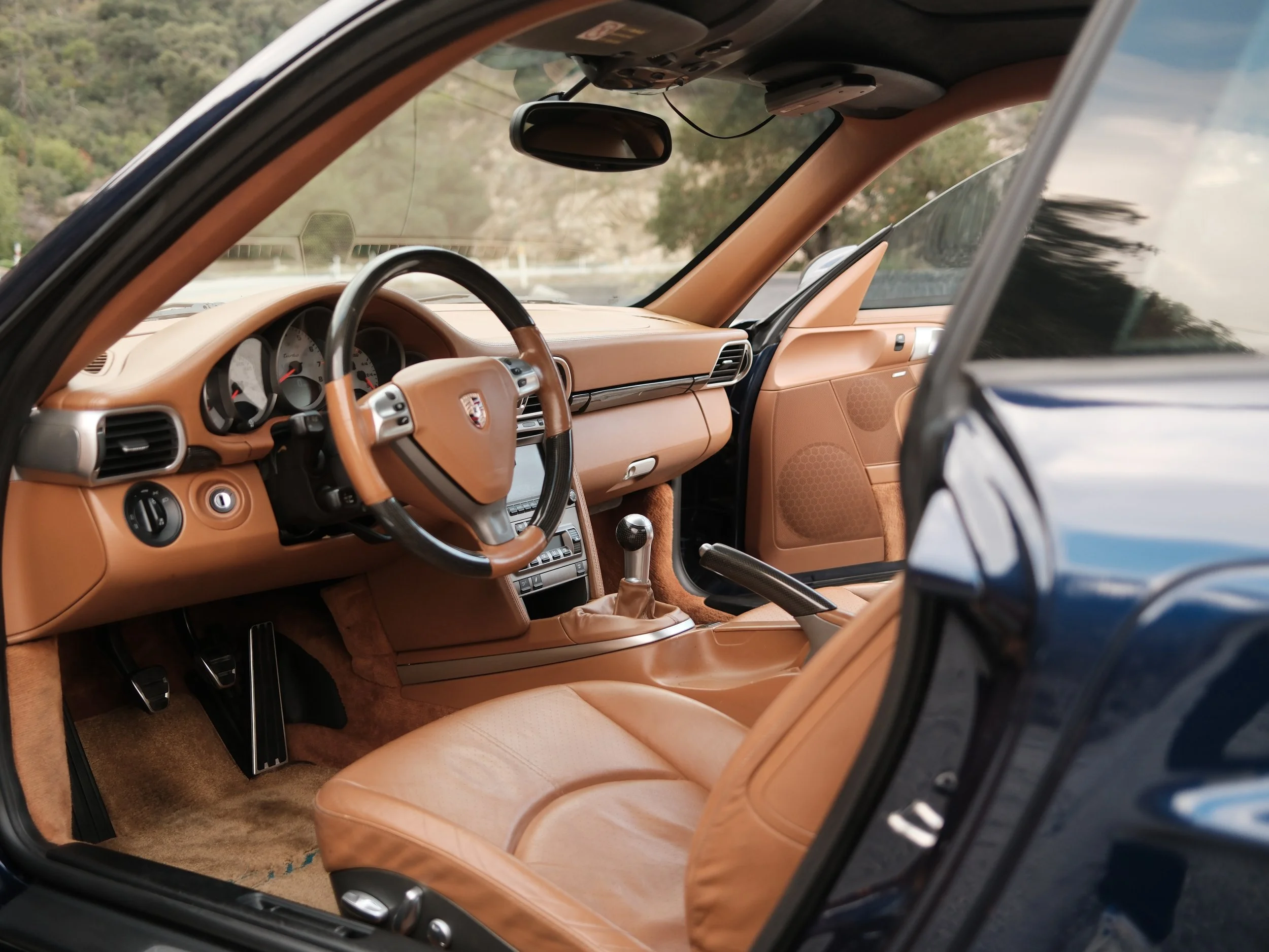 Interior of a classic Porsche sports car with tan leather seats and dashboard, featuring a sport steering wheel and manual gear shift.