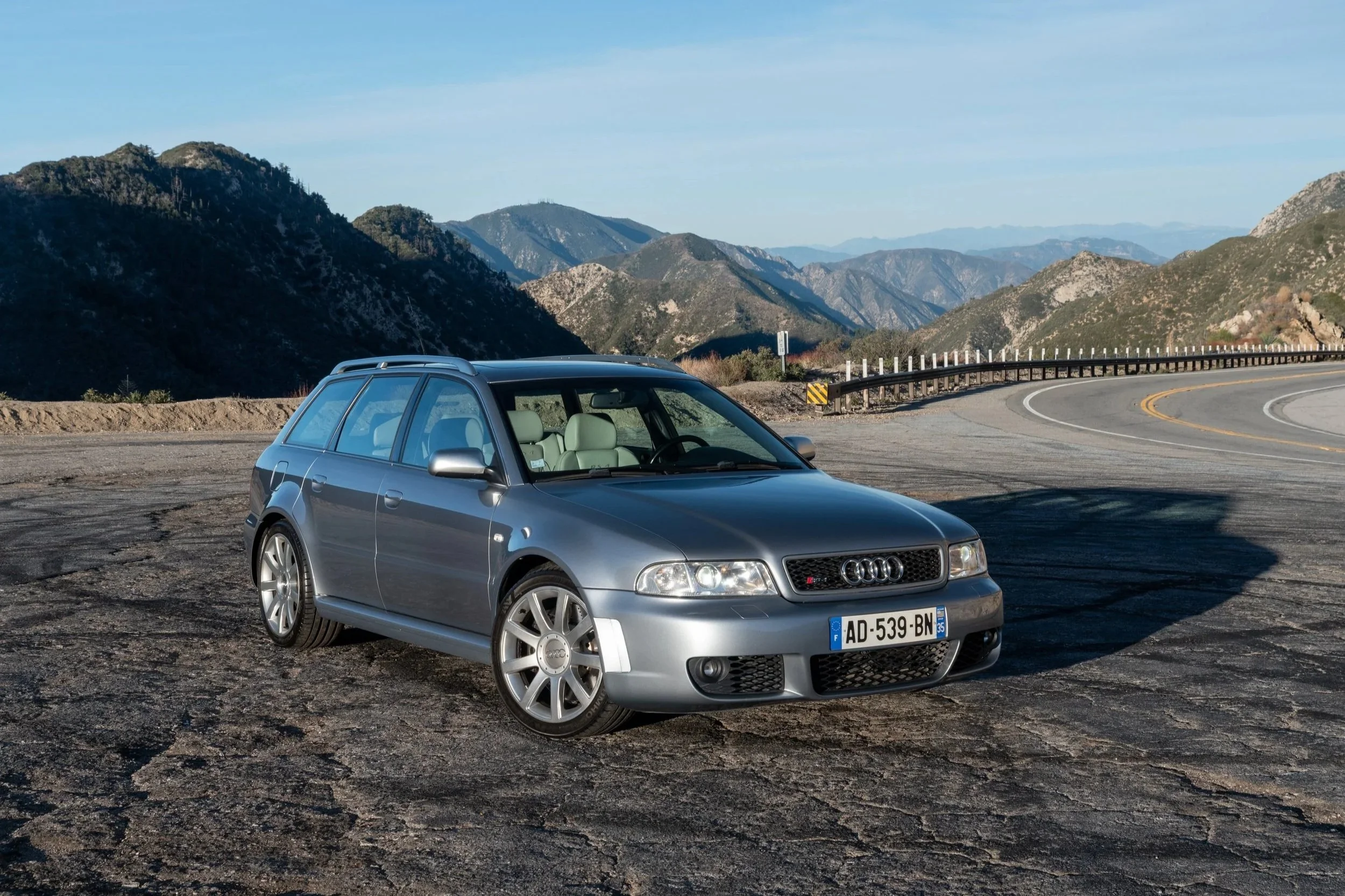 A silver Audi station wagon parked on a dirt and gravel area beside a mountain road, with mountain ranges in the background and a clear blue sky.