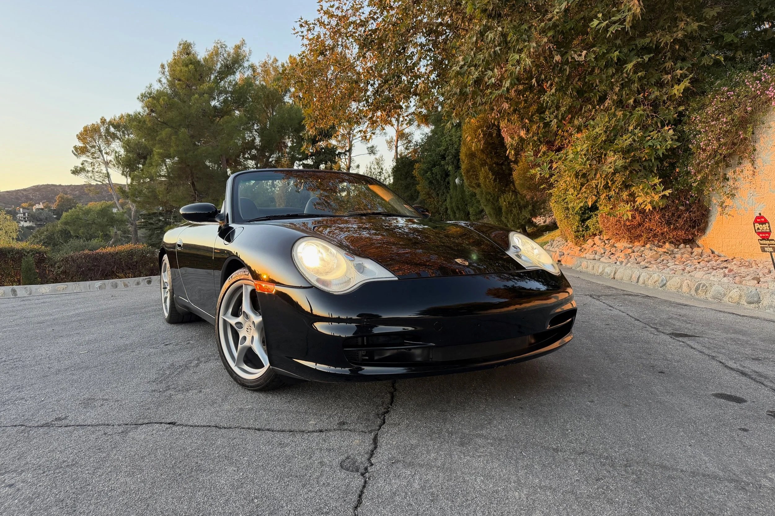 A black Porsche convertible car parked on a paved surface surrounded by trees and bushes during sunset.