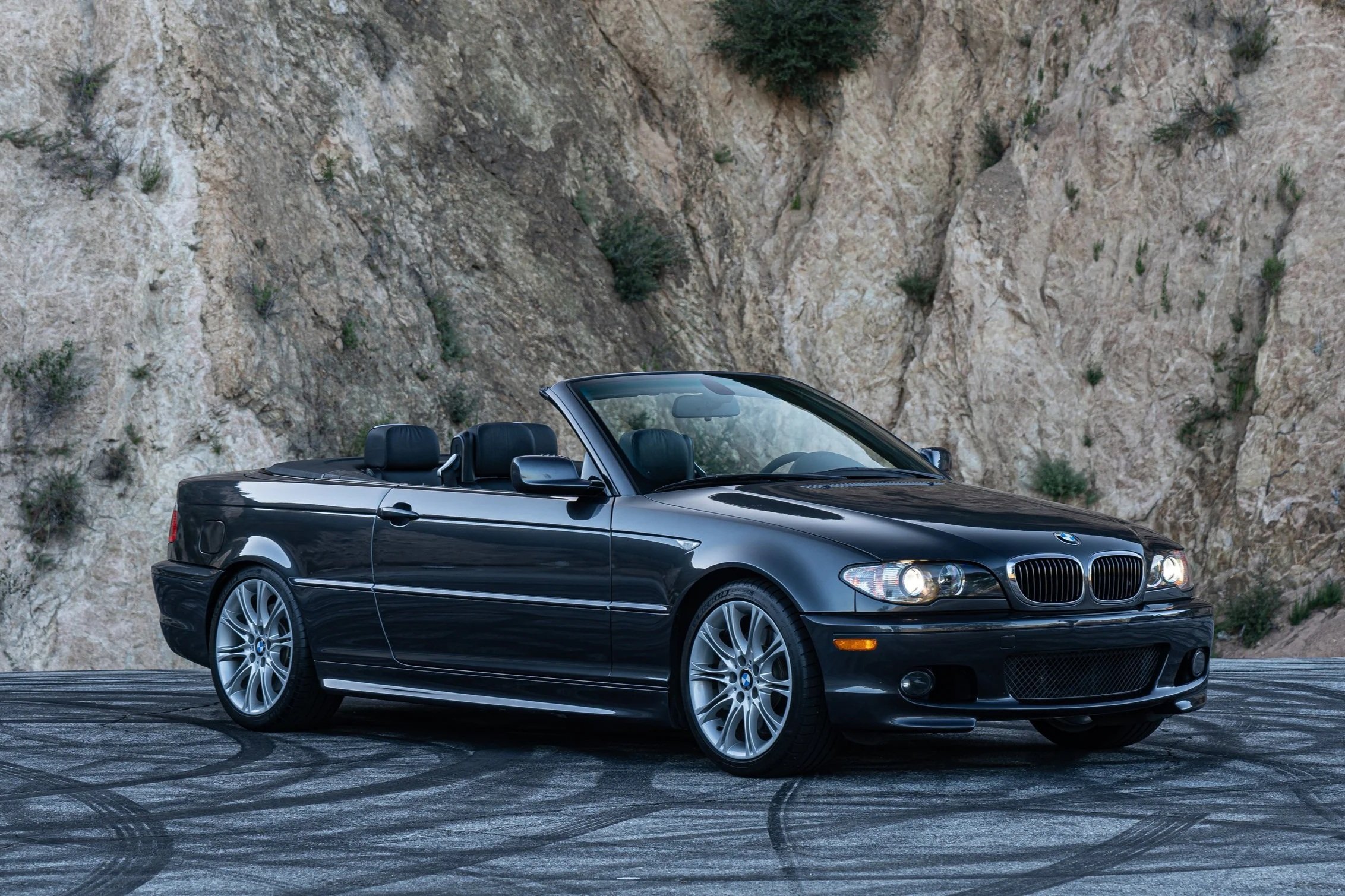 A black BMW convertible parked on a paved surface with tire marks, against a rocky hillside background.