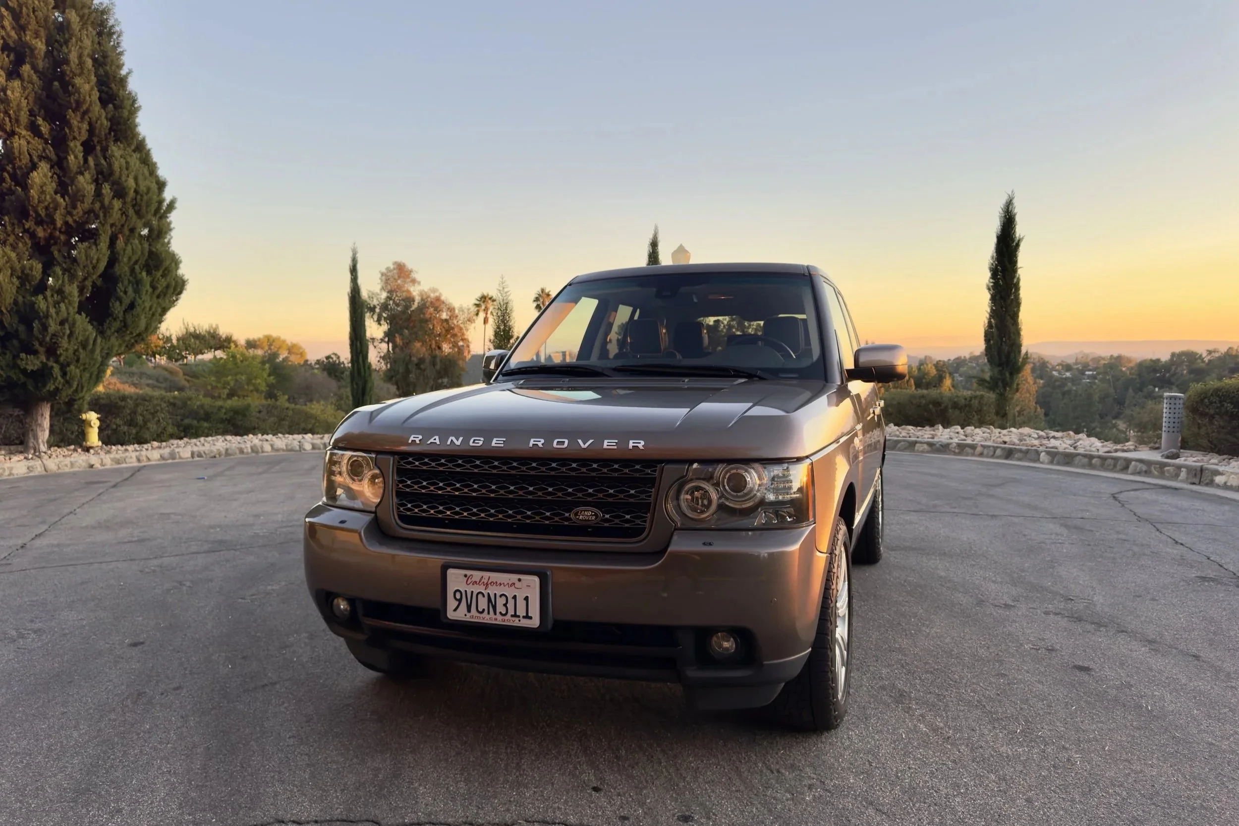 A gray Range Rover SUV parked on a driveway with trees and bushes in the background during sunset.