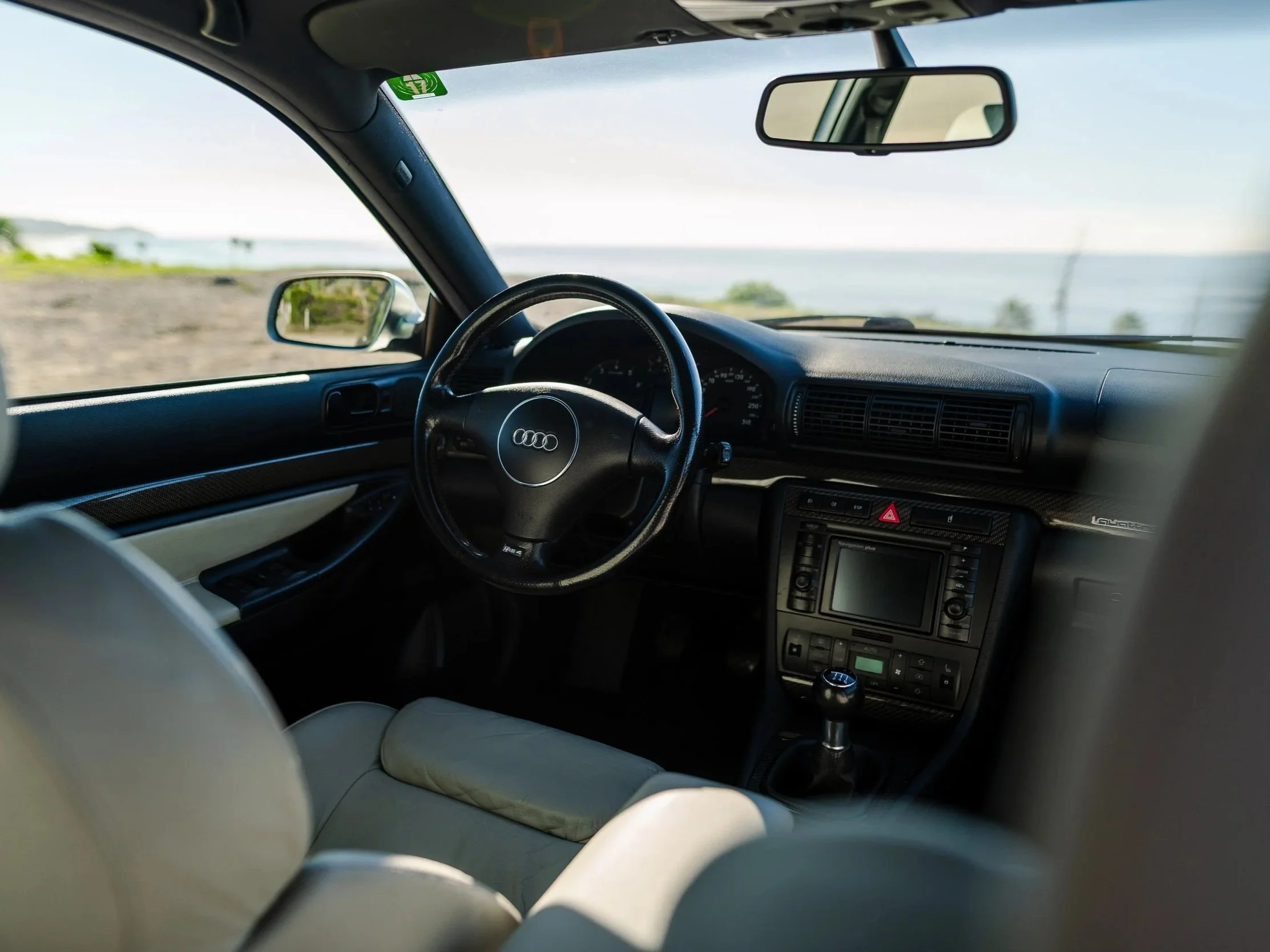 Interior of an Audi car parked near a body of water, showing the dashboard, steering wheel, and front seats with beige upholstery, with a view of a beach and ocean through the windshield.