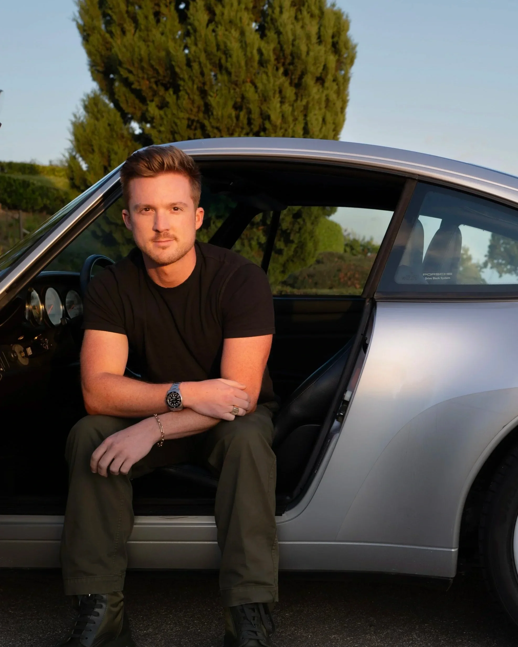 A young man sitting on the door sill of a silver sports car with the driver's door open, outdoors during sunset, with trees in the background.
