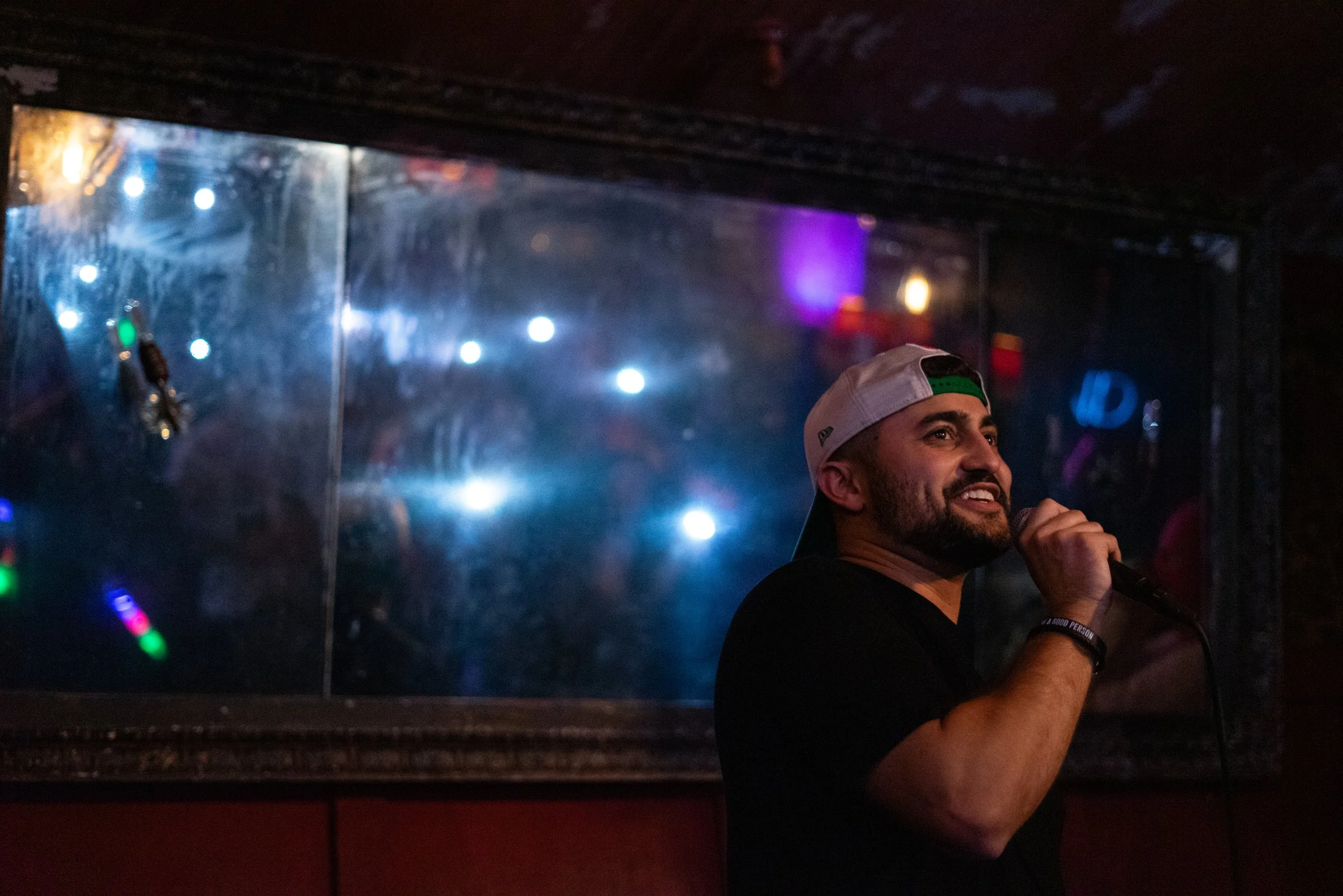 Nadroj performing wearing a black shirt and a white Philadelphia Eagles cap backward is holding a microphone and smiling, with a dark, colorful, illuminated background behind a window or glass panel. Nadroj musical artist comedy rapper.