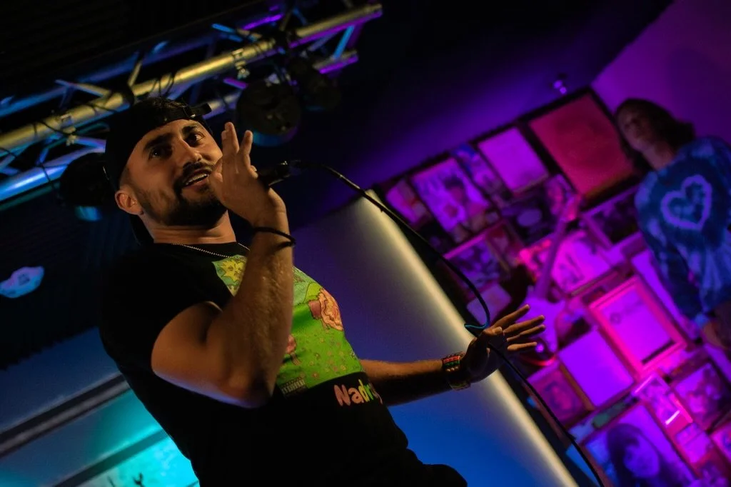 Nadroj holding a microphone, wearing a black cap and a black T-shirt with a colorful graphic, performing on stage with a background of vibrant purple and pink lights and people in the audience. Nadroj musical artist comedy rapper.