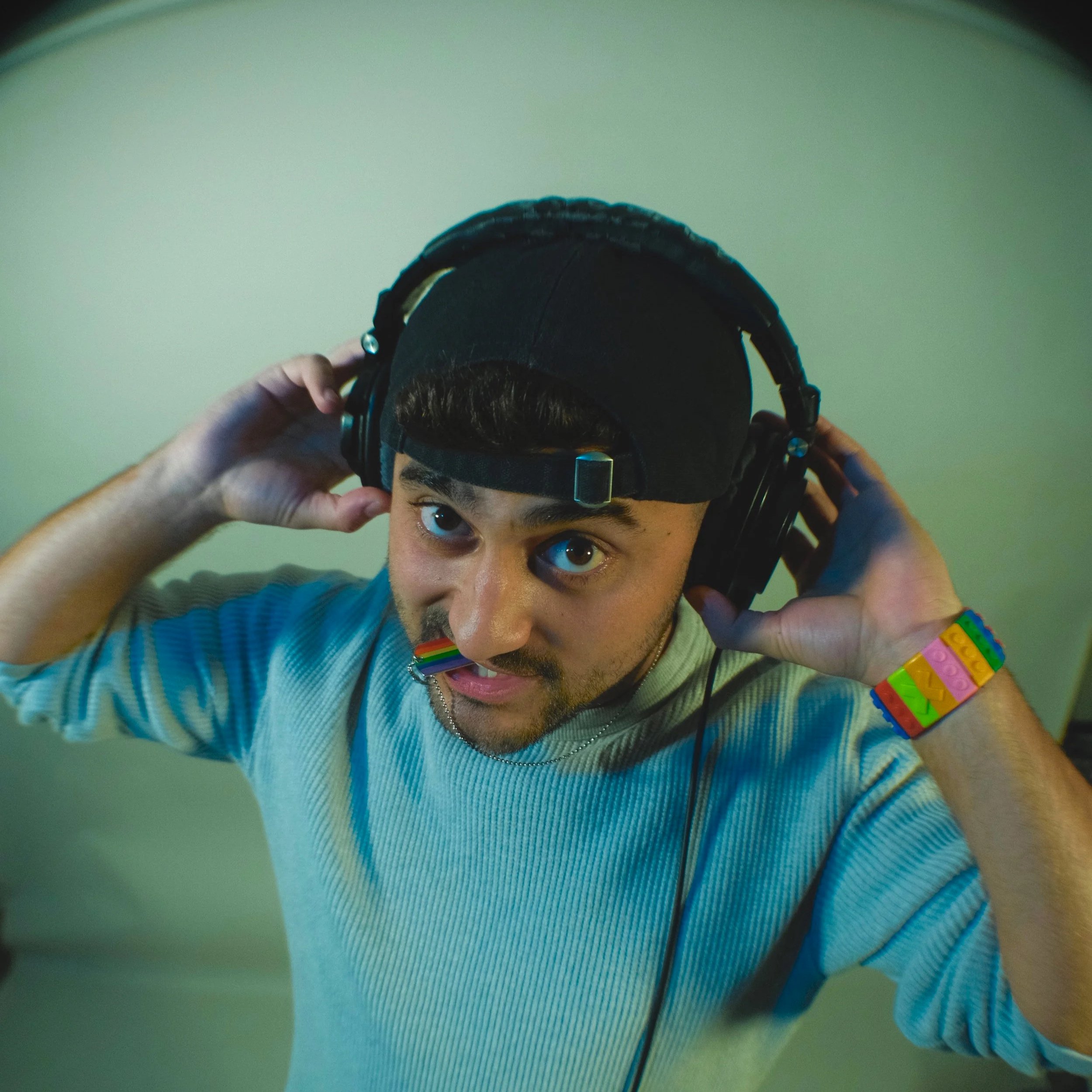 Nadroj wearing a rainbow-colored bracelet and a rainbow-colored dog tag, wearing headphones and a black cap, adjusting the headphones while looking up at the camera. Nadroj musical artist comedy rapper.