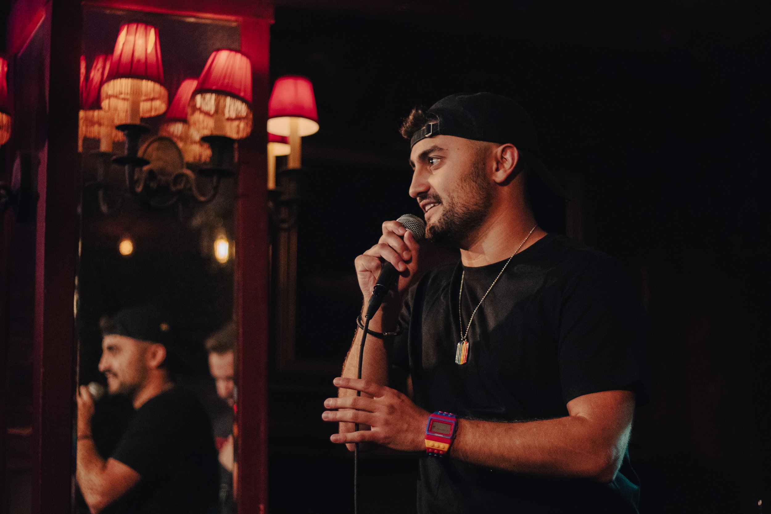 Nadroj holding a microphone on stage in a dimly lit venue with red lamps, wearing a black cap backwards, black t-shirt, rainbow-colored watch, and rainbow necklace, smiling. Nadroj musical artist comedy rapper.