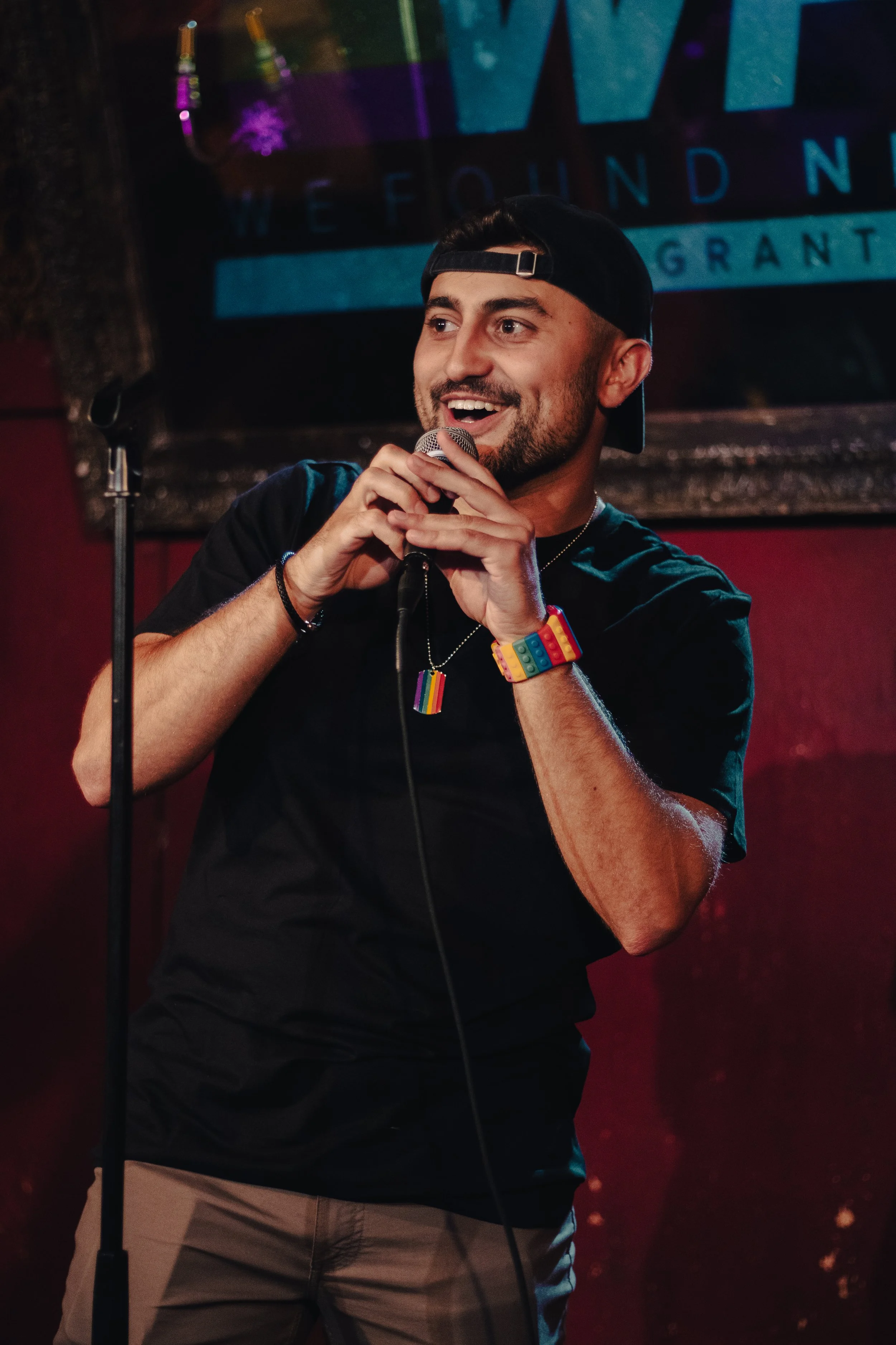 Nadroj wearing a black cap backwards, a black T-shirt, and rainbow pride accessories, smiling and holding a microphone on stage at a concert. Nadroj musical artist comedy rapper.