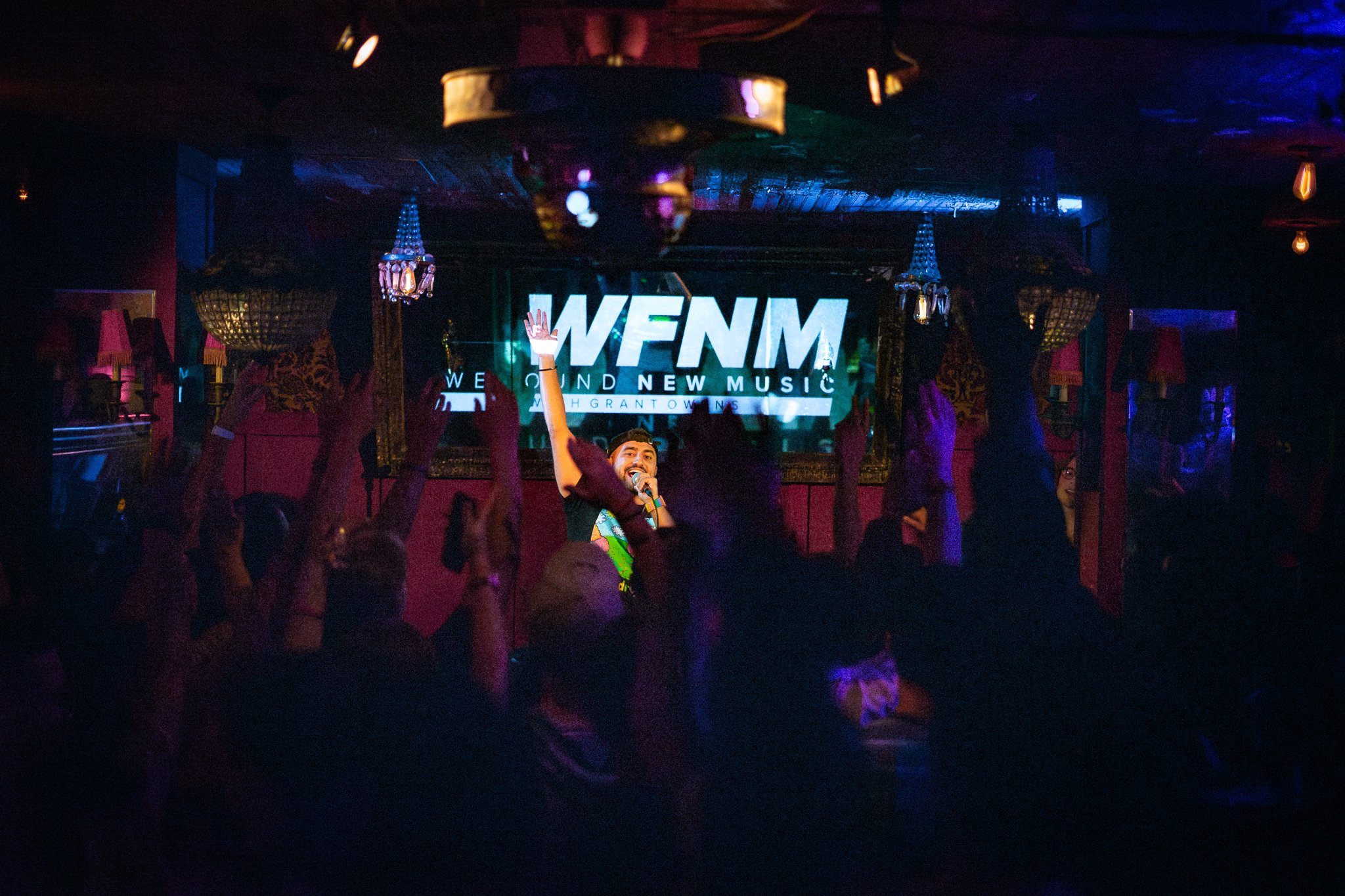 Nadroj performing and rapping with a microphone on stage at an event, with the WFNM radio logo on a screen behind him and an audience raising their hands. Nadroj musical artist comedy rapper. Bar Lubitsch, West Hollywood.
