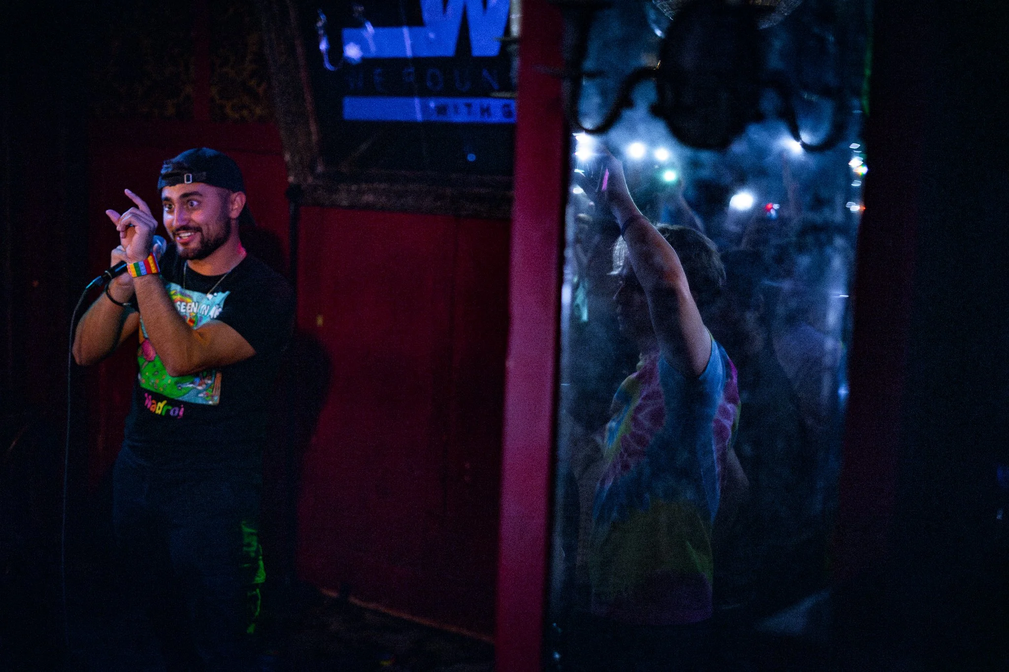 Nadroj on stage smiling and speaking into a microphone, wearing a black cap and a tie-dye wristband in a dimly lit venue. Beside him a person behind a reflective surface is partially visible raising their hand, with colorful lights in the background.