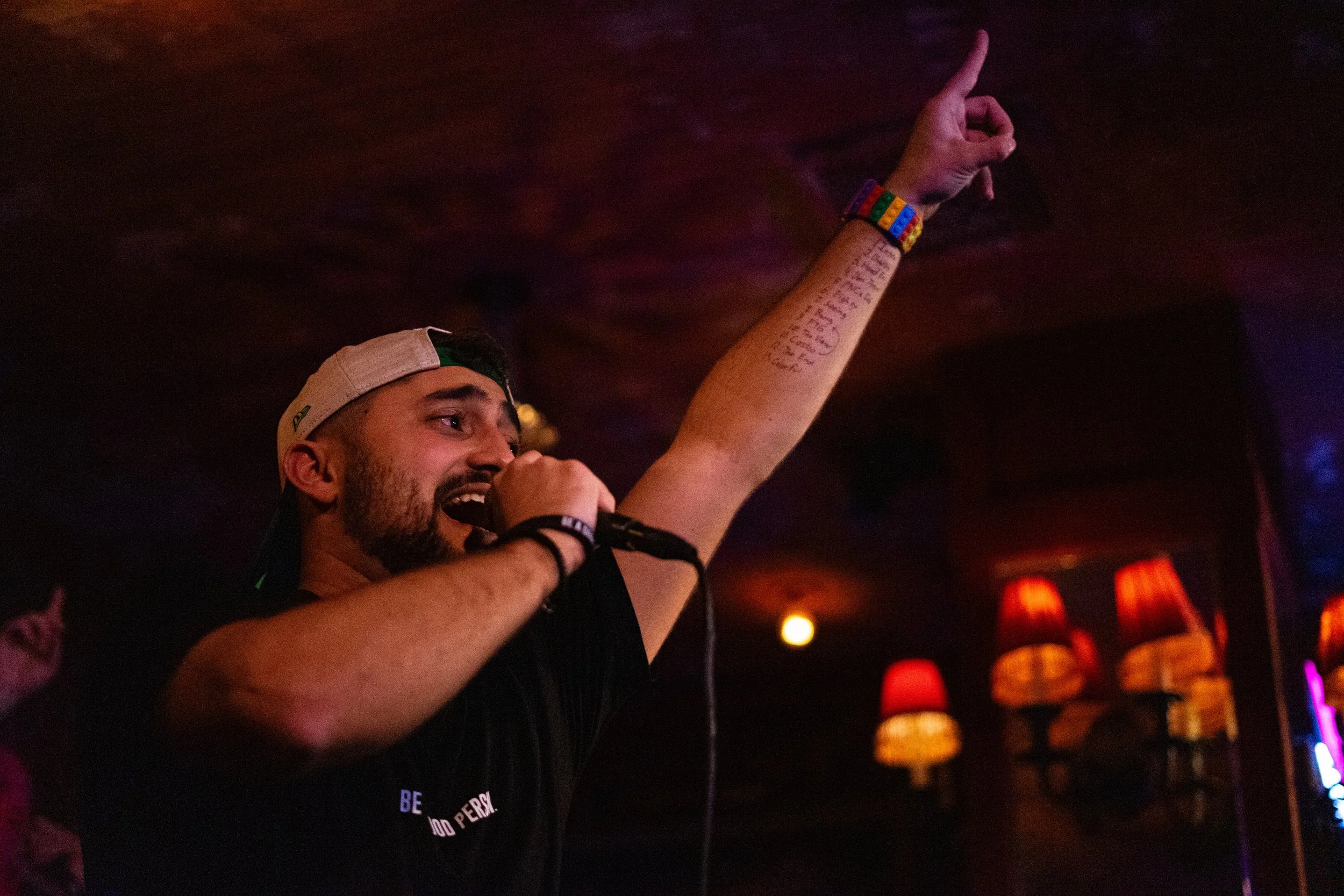 Nadroj wearing a backwards baseball cap, singing into a microphone, with his right arm raised and pointing upward. He has a setlist on his forearm and is wearing a colorful rainbow bracelet. Nadroj musical artist comedy rapper.