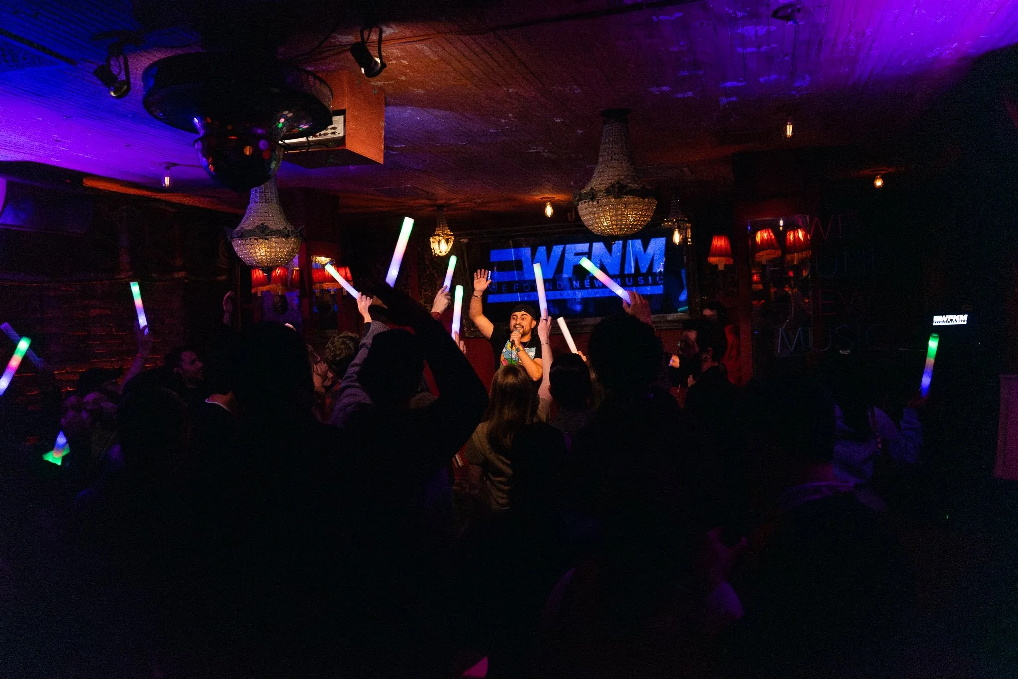 Nadroj performing on stage at a dimly lit nightclub, holding a microphone and waving, with an audience holding colorful glow sticks. The background features a large screen displaying 'WFNM' and chandeliers hanging from the ceiling.