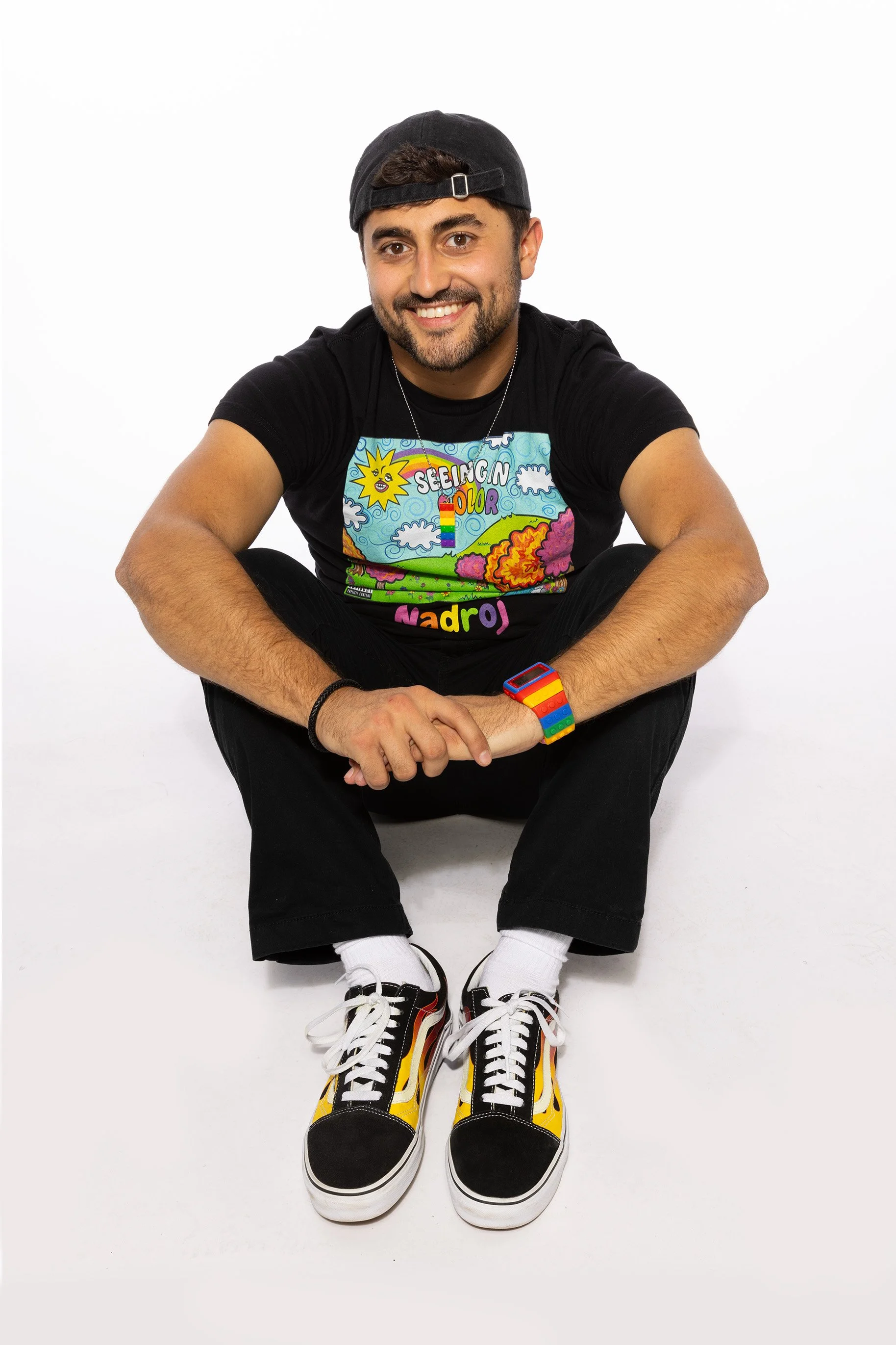 Nadroj smiling, sitting cross-legged on a white background, wearing a black cap, colorful sneakers, black pants, a colorful watch, and a black t-shirt with his Seeing in Color mixtape artwork on it - vibrant graphics and text.