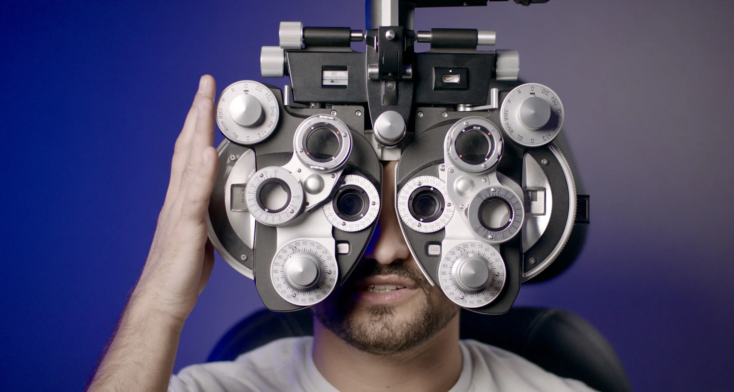 A person wearing a trial frame with multiple adjustable lenses, commonly used by optometrists for eye examinations, against a gradient blue background. Nadroj Seeing in Color musical artist comedy rapper.