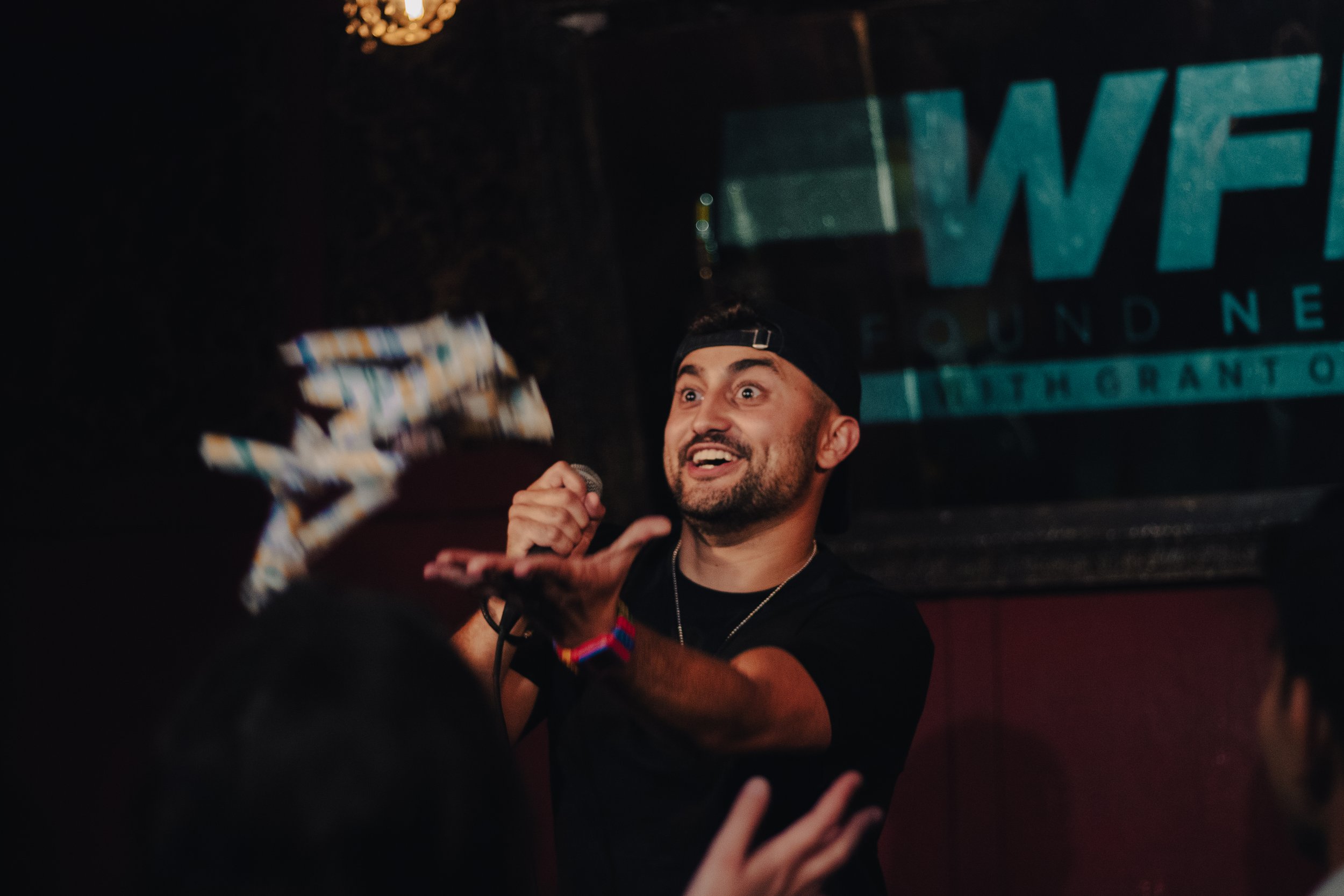 Nadroj in a black cap holding a microphone, smiling and engaging with an audience in a dimly lit space with a sign in the background. Nadroj musical artist comedy rapper.
