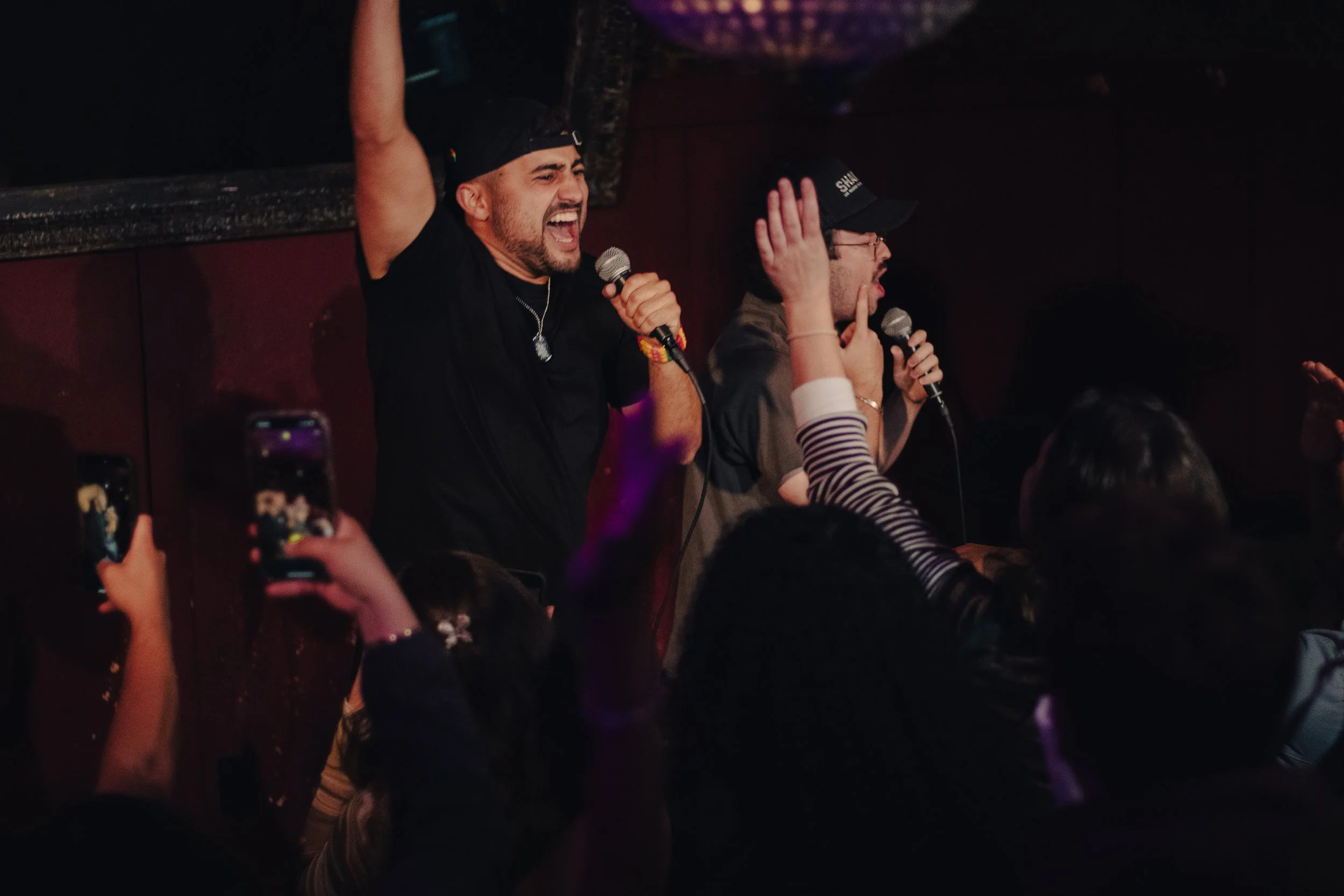 Nadroj singing on stage with DJ microphones, one with a black cap and the other with a black cap and glasses, crowd in the foreground with some people raising hands and taking photos. Nadroj musical artist comedy rapper.