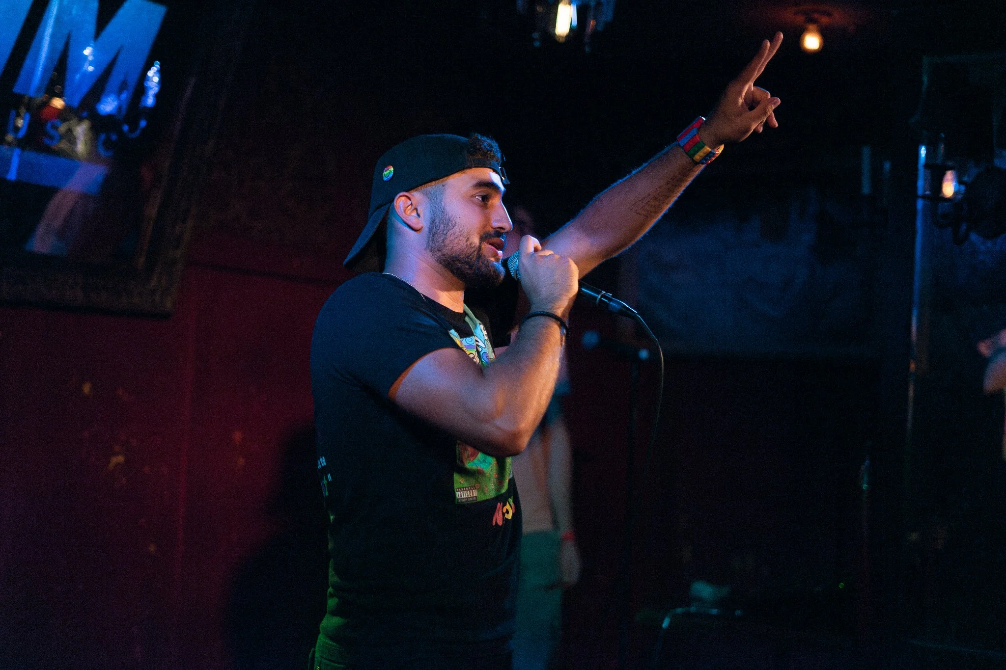 Nadroj wearing a black T-shirt and a backward baseball cap singing into a microphone with one arm raised, performing in a dimly lit venue. Nadroj musical artist comedy rapper performing.