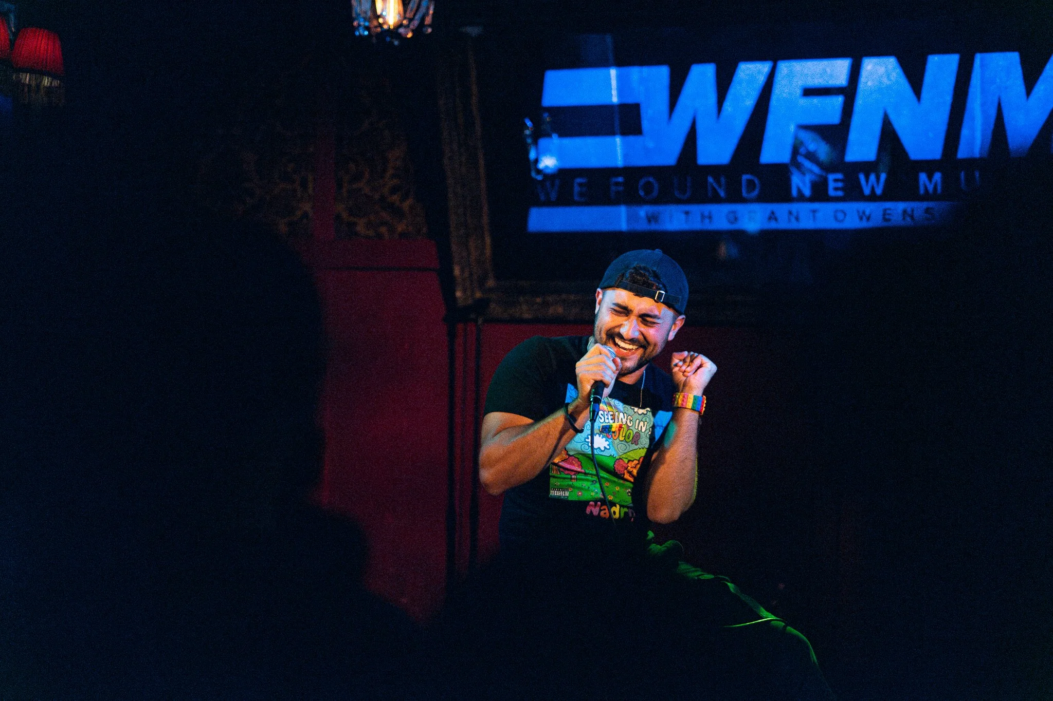 NADROJ singing or speaking into a microphone with a big smile, seated in a dark venue, with a blue screen in the background displaying the text 'WFNM WE FOUND NEW MUSIC'. Nadroj musical artist comedy rapper.