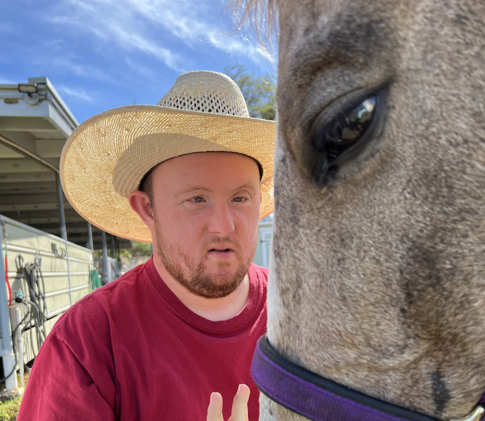A man with reddish hair and a beard wearing a large straw sun hat and red T-shirt, standing next to a large horse with gray fur and a purple halter, in an outdoor setting with a blue sky and some farm equipment.