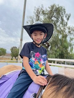 Young boy wearing a black cowboy hat and a graphic t-shirt, smiling while riding a horse outdoors with trees and a cloudy sky in the background.