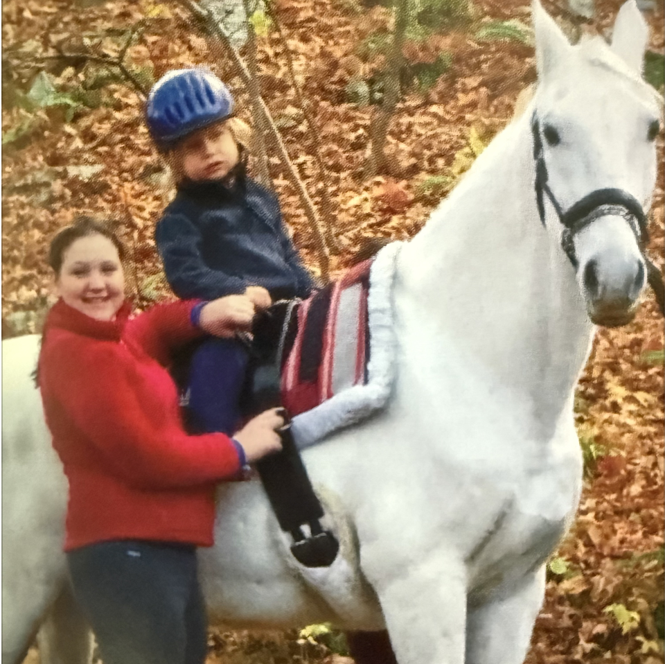 A young boy wearing a blue helmet riding a white horse with a woman standing beside in an autumn forest setting.