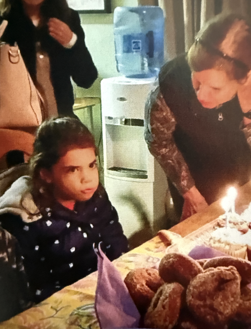 Girl with dark hair and polka dot jacket at birthday celebration with cake, candles, and family members in indoor setting.